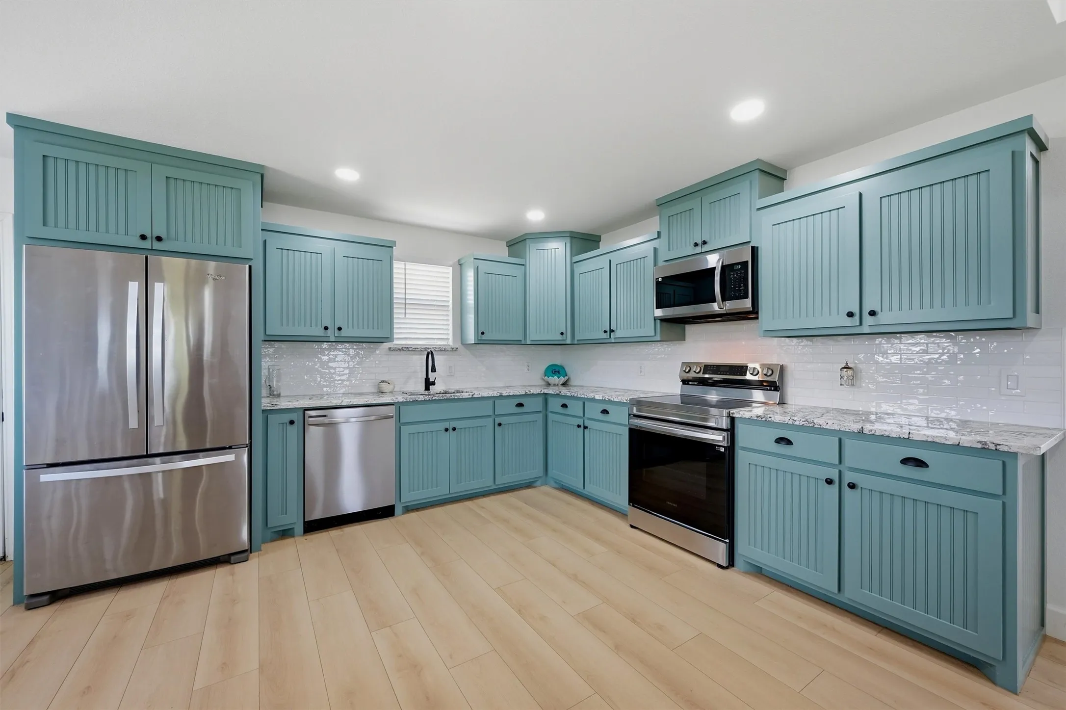 Kitchen featuring light wood-finish flooring, custom cabinetry with beadboard paneling, stainless steel appliances, white subway tile backsplash, and light-toned countertops