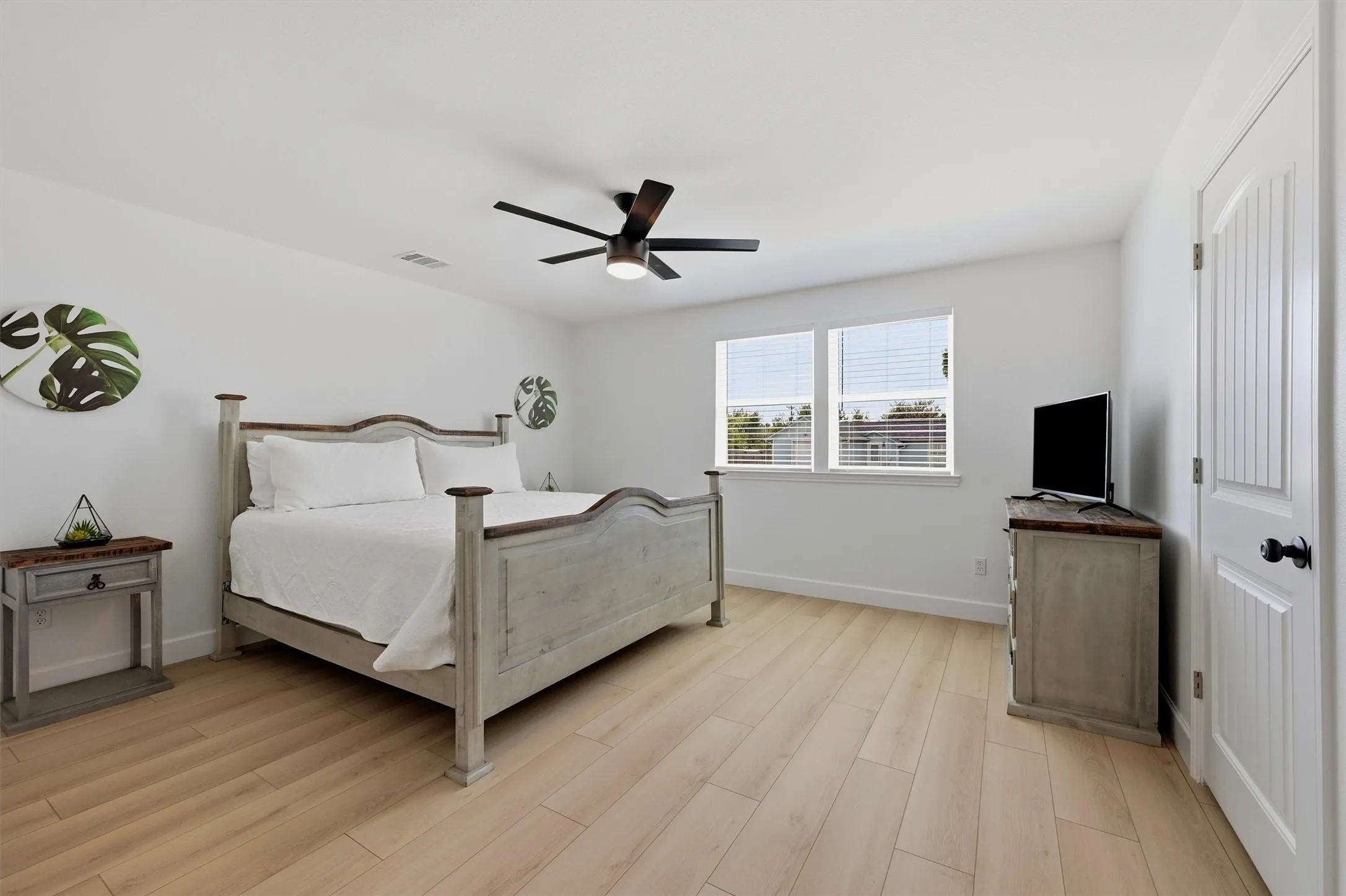 Light wood-finish flooring, dual windows with blinds, ceiling fan with integrated lighting, white interior door with dark hardware, and neutral wall paint
