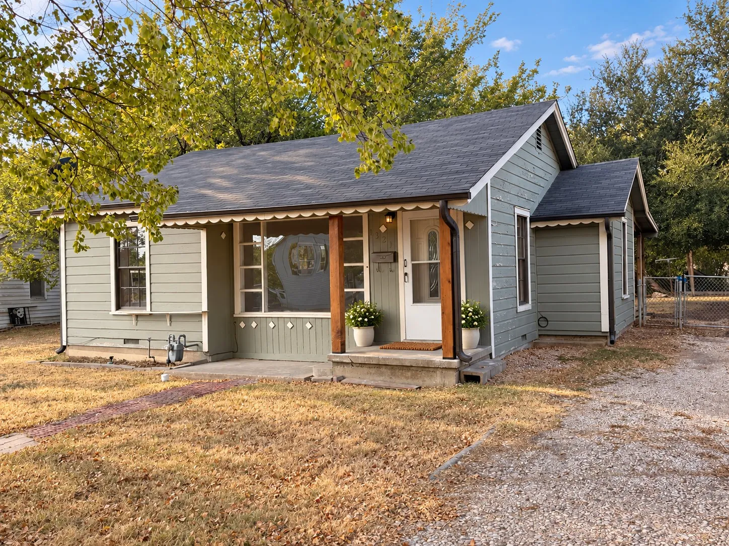 Light green siding with white trim, dark shingle roof, decorative scalloped eaves, covered porch with wood support beam, and gravel driveway