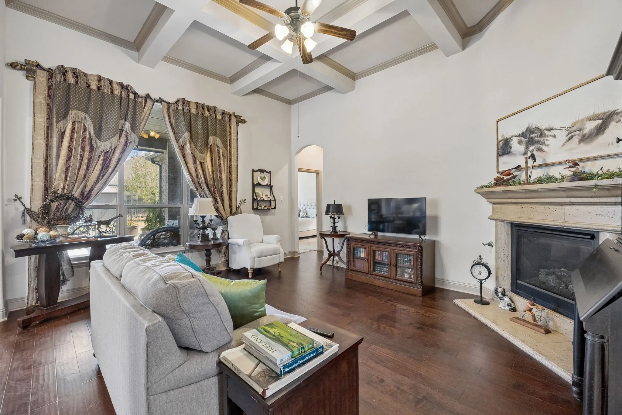 Tray ceiling with exposed beams, wood-finish flooring, stone-clad fireplace with mantel, arched doorway, and large windows