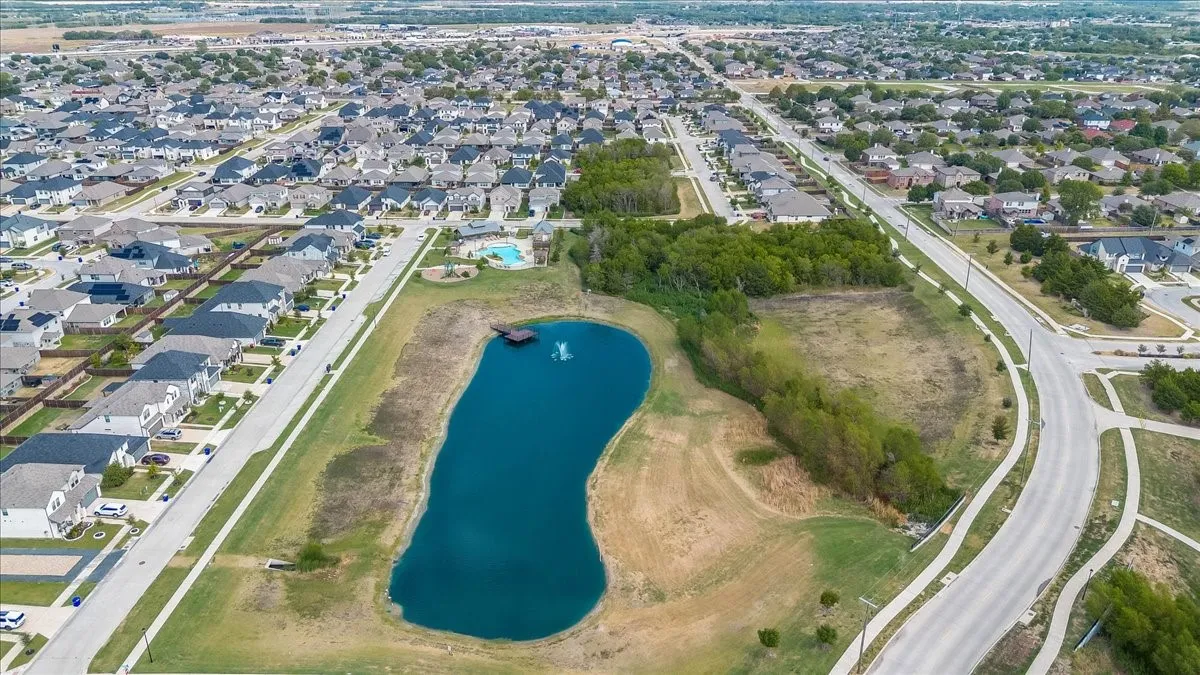 Aerial perspective of a developed residential community featuring numerous single-family homes, a central pond with a water feature and dock, a community swimming pool, and paved streets with sidewalks