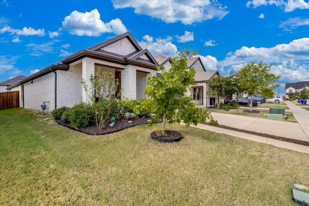 White brick exterior with dark trim accents, covered front porch, landscaped garden beds, front lawn, and concrete sidewalk