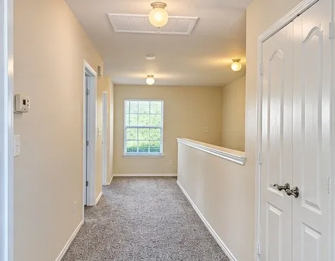 Neutral wall color, carpeted hallway, multiple ceiling-mounted light fixtures, window with grid pattern, white interior doors