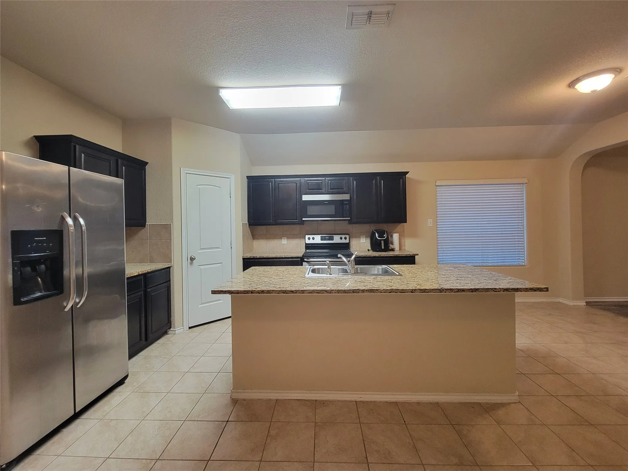 Kitchen featuring dark wood-finish cabinetry, stainless steel appliances, a center island with an integrated sink, tile flooring, and light-colored countertops