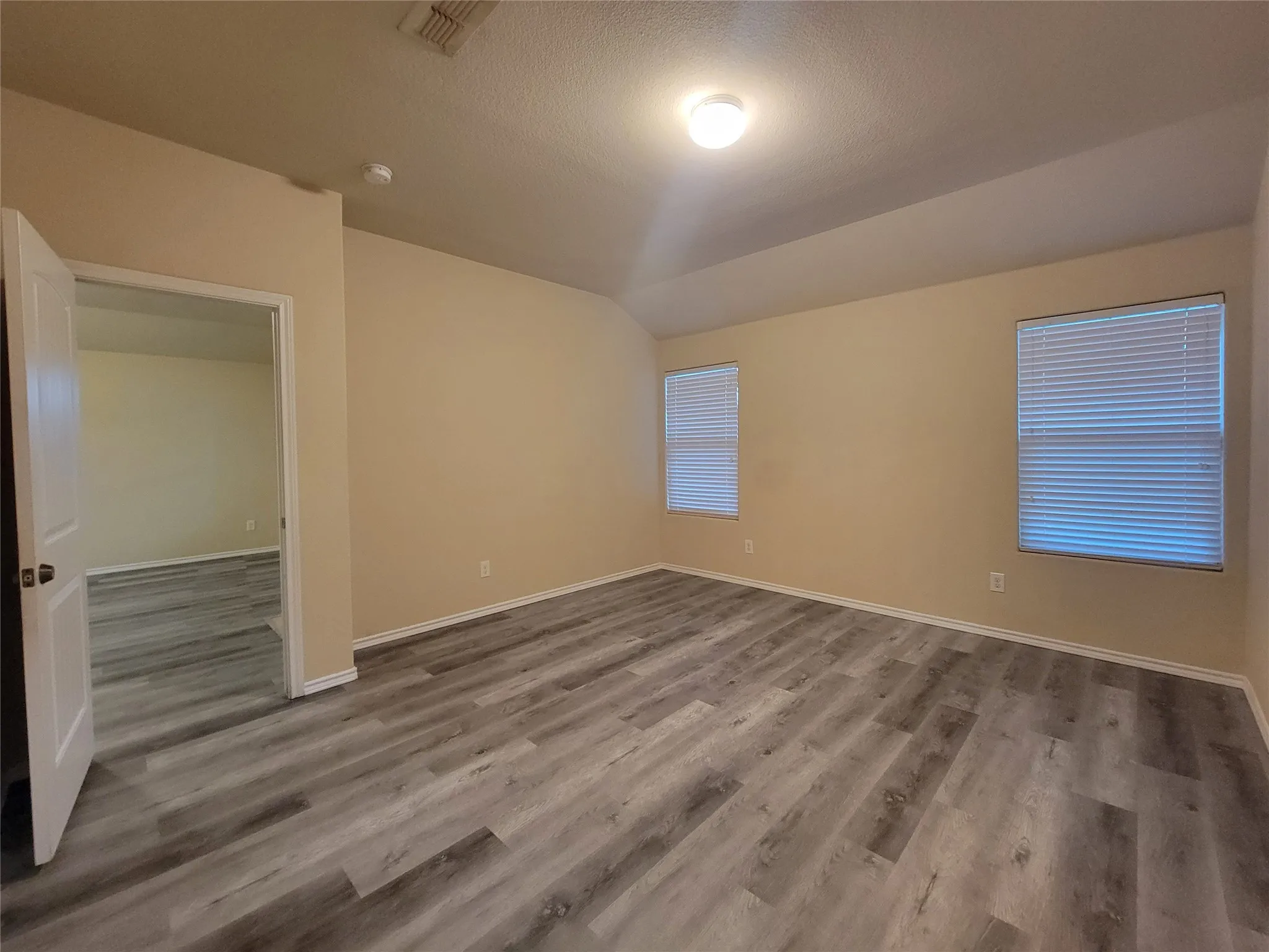 Spacious room featuring wood-finish flooring, two windows with blinds, and an overhead light fixture