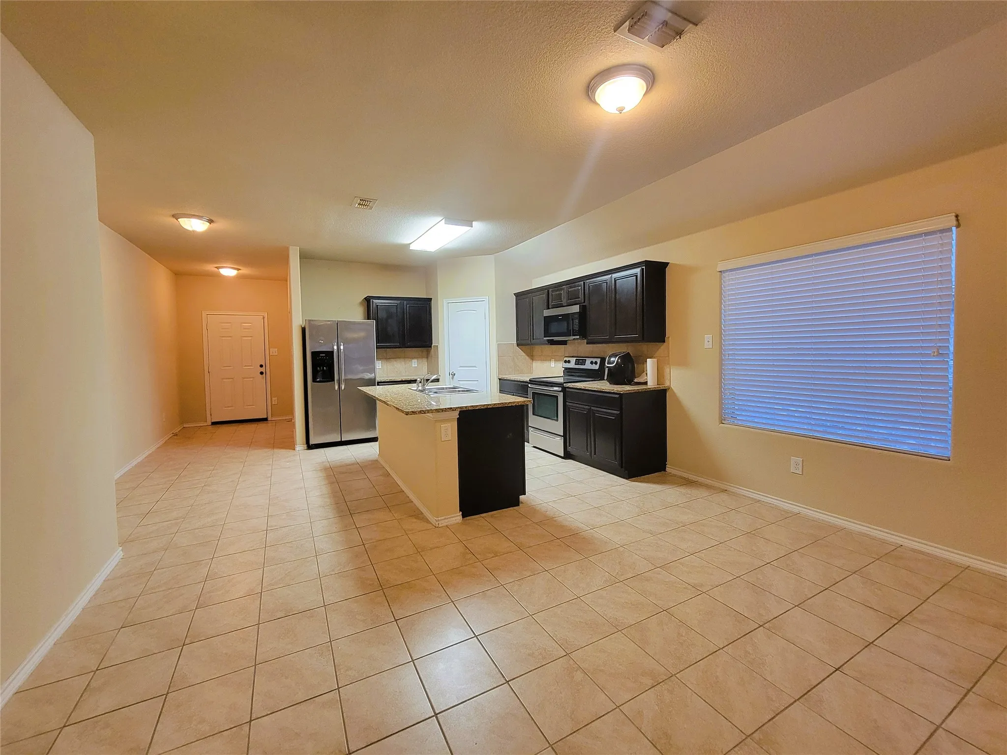 Open-concept living space with tile flooring, dark wood-finish cabinetry, stainless steel appliances, and a kitchen island with a stone-finish countertop