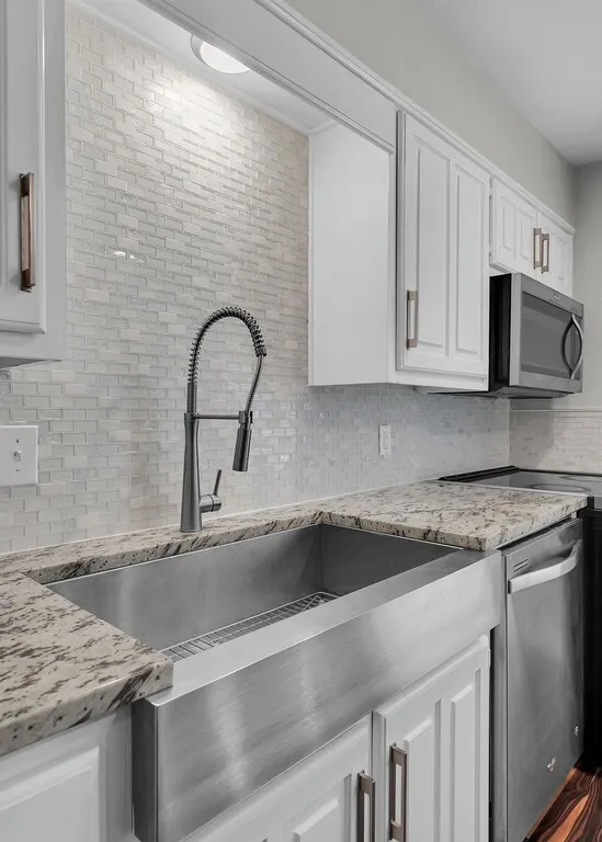 Stainless steel farmhouse sink with a gooseneck faucet, light-toned granite countertops, white shaker-style cabinetry, and a subway tile backsplash