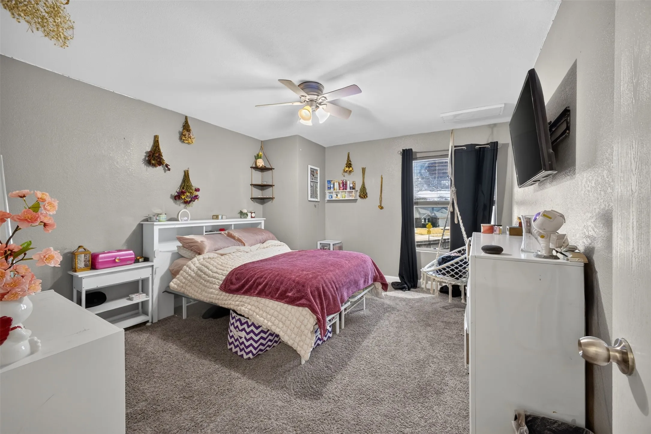 Plush carpeting, ceiling fan with integrated lighting, window with dark drapes, wall-mounted shelving, and neutral wall paint