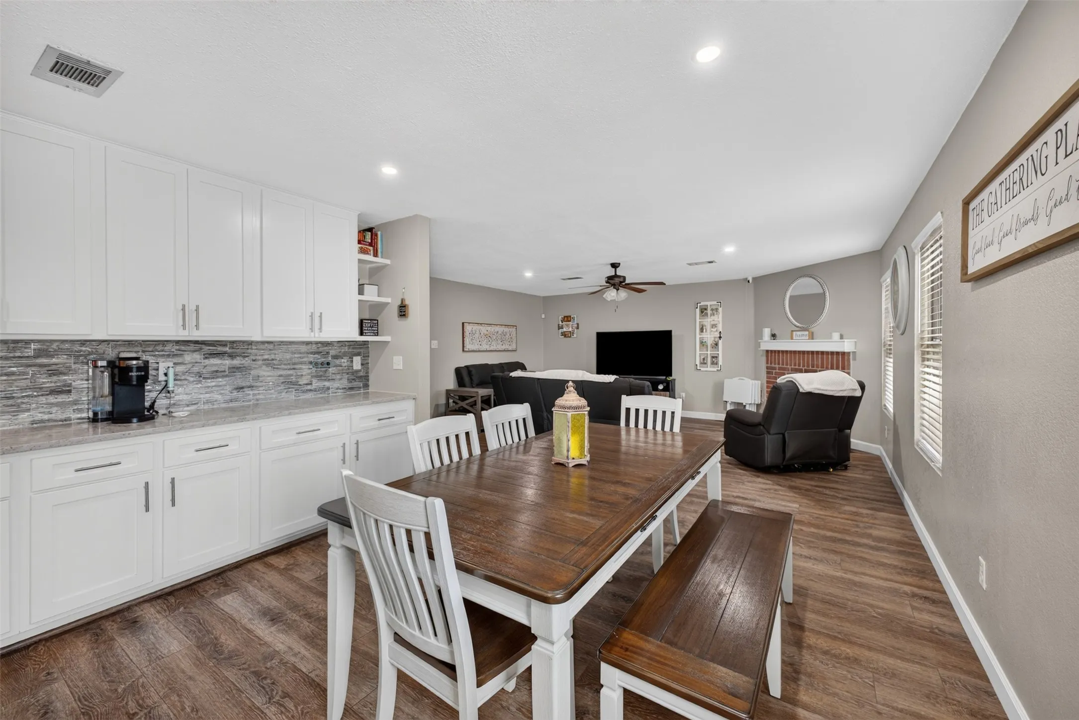 White cabinetry with modern hardware, stone-look backsplash, wood-finish flooring, recessed lighting, and a brick-front fireplace
