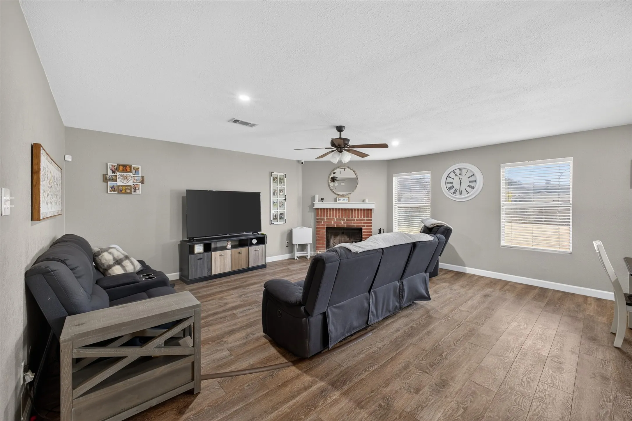 Open-concept living area with wood-finish flooring, a brick-surround fireplace, and a ceiling fan with integrated lighting