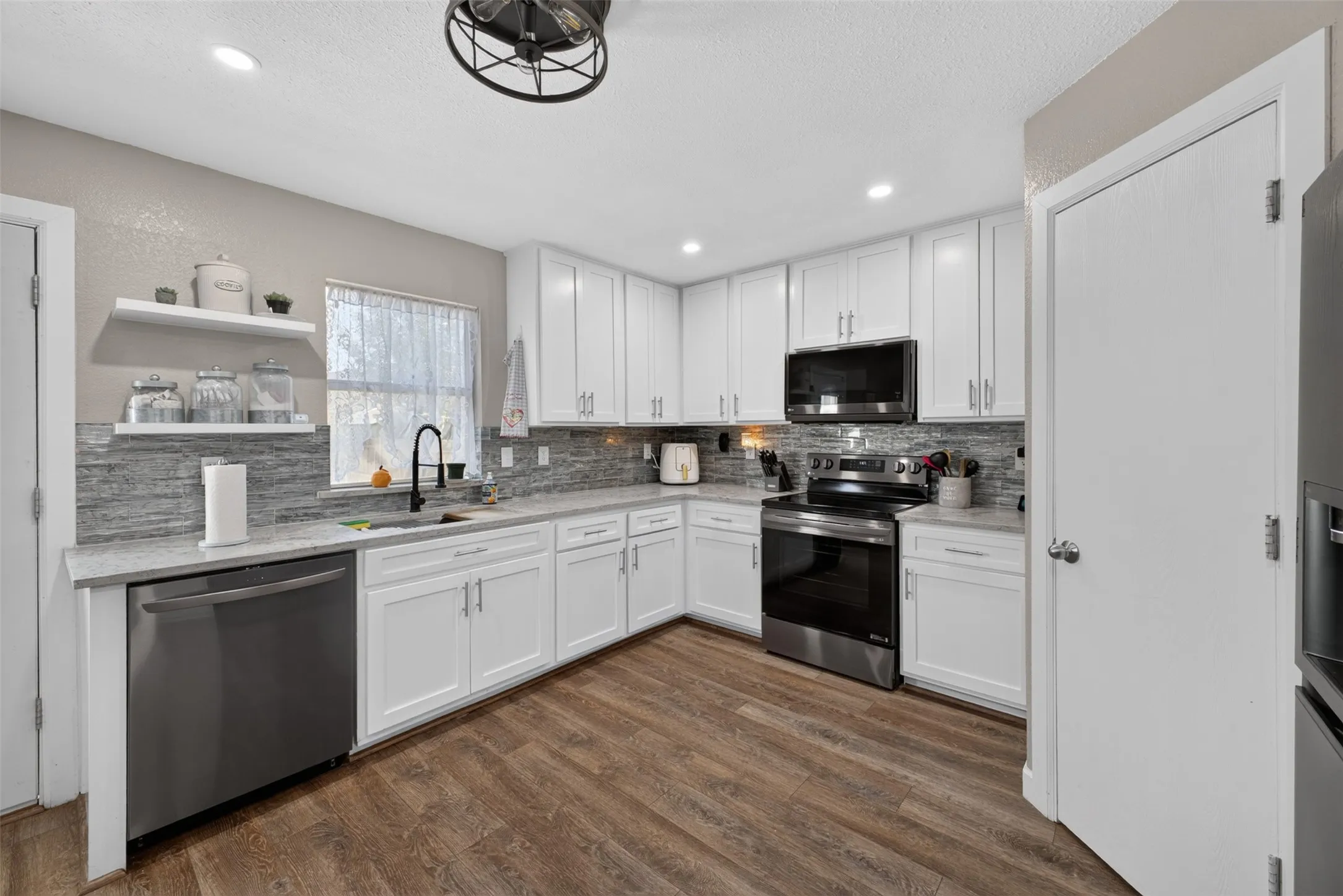 Kitchen with wood-finish flooring, white cabinetry, integrated stainless steel appliances, light-toned countertops, and a full-height tile backsplash