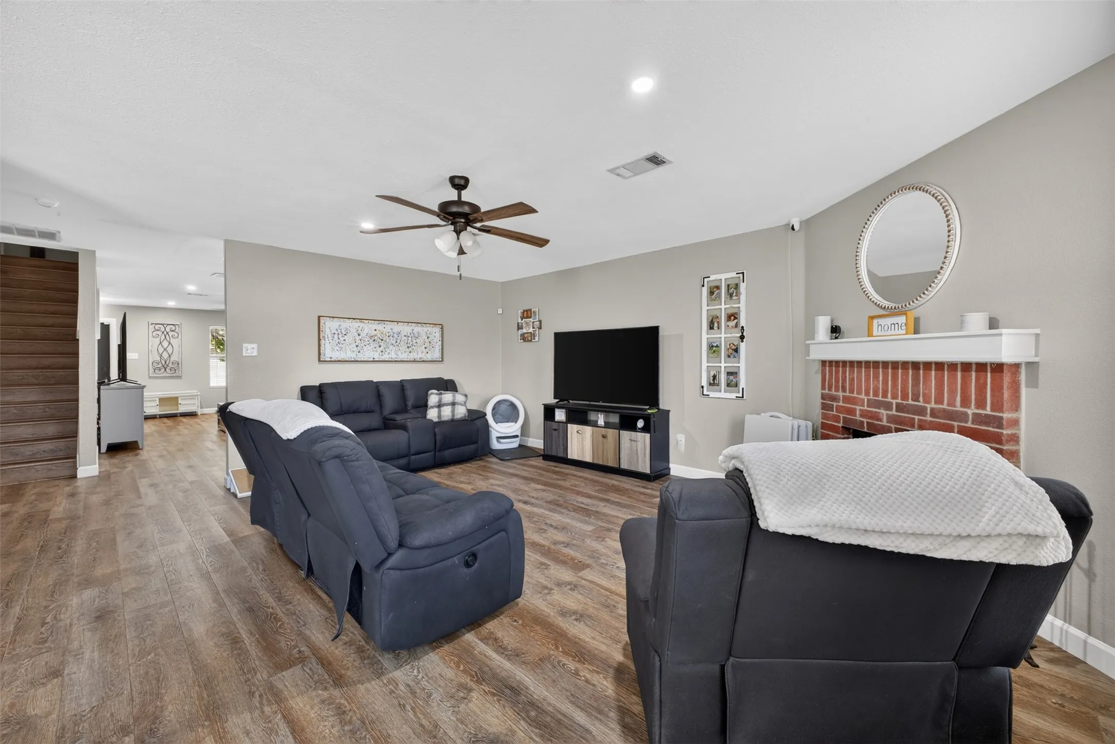 Wood-finish flooring, brick fireplace with white mantel, ceiling fan with light fixture, and neutral wall tones