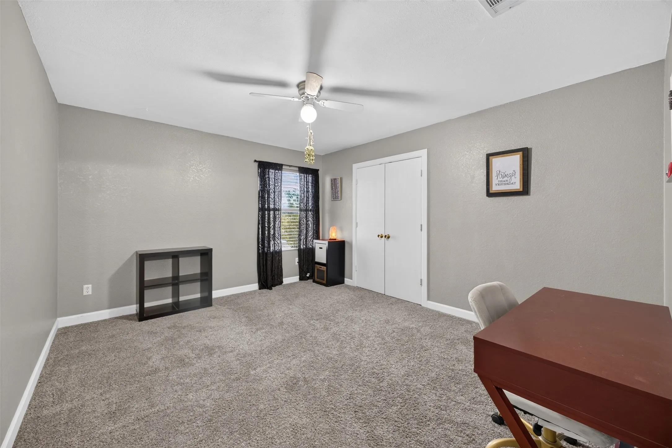 Gray textured walls, neutral gray carpeting, white ceiling with a ceiling fan, white bi-fold closet doors, and a window with dark window treatments