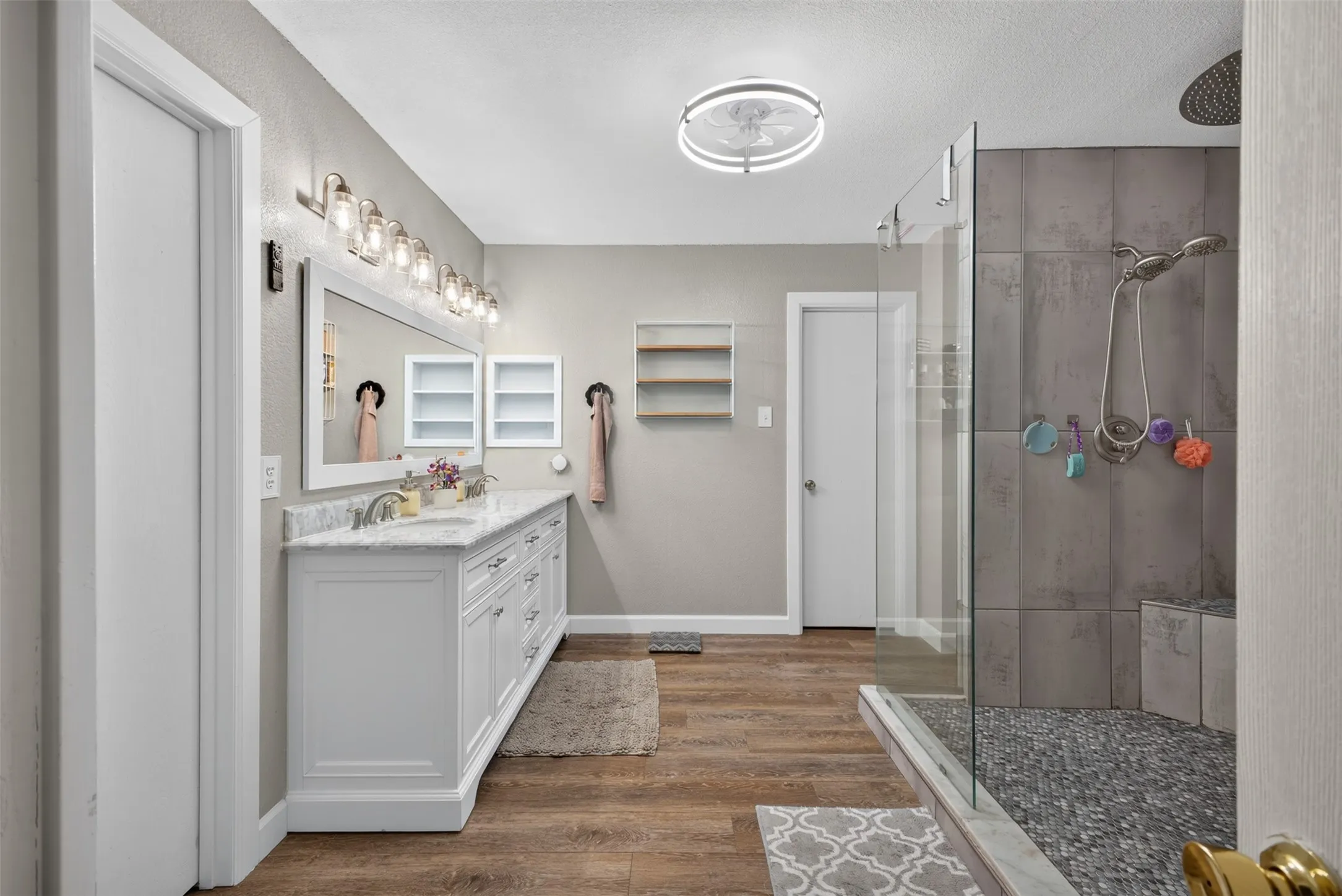 Spacious bathroom featuring a double vanity with white cabinetry, a large mirror, a multi-bulb vanity light, wood-finish flooring, and a walk-in shower with glass enclosure and mosaic tile base