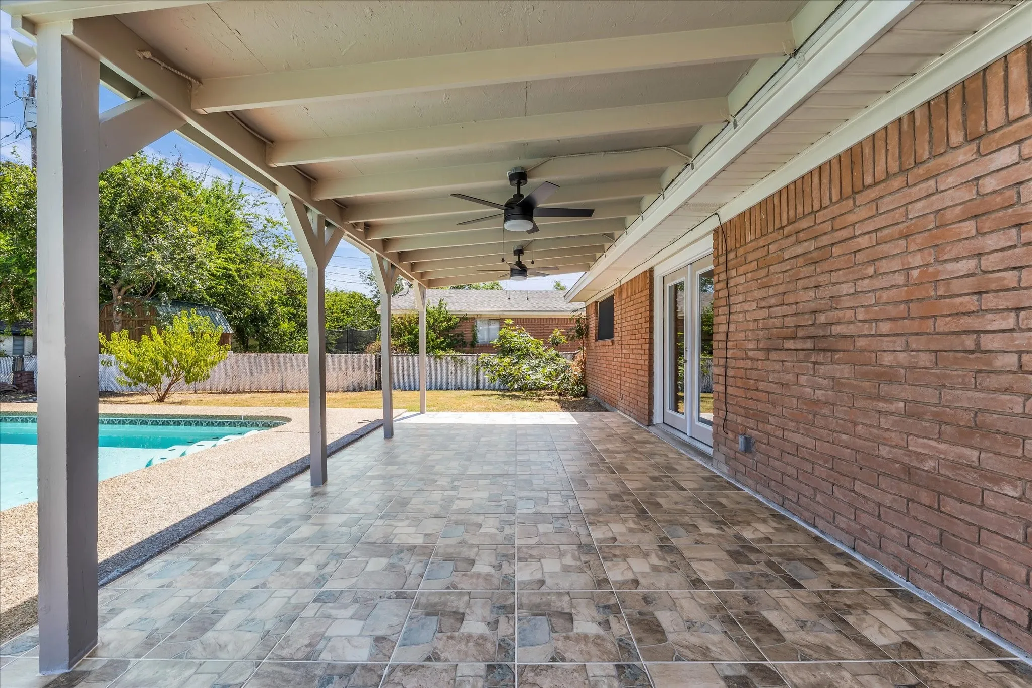 Spacious covered patio with ceiling fans, tiled flooring, brick exterior, and a swimming pool with surrounding hardscaping