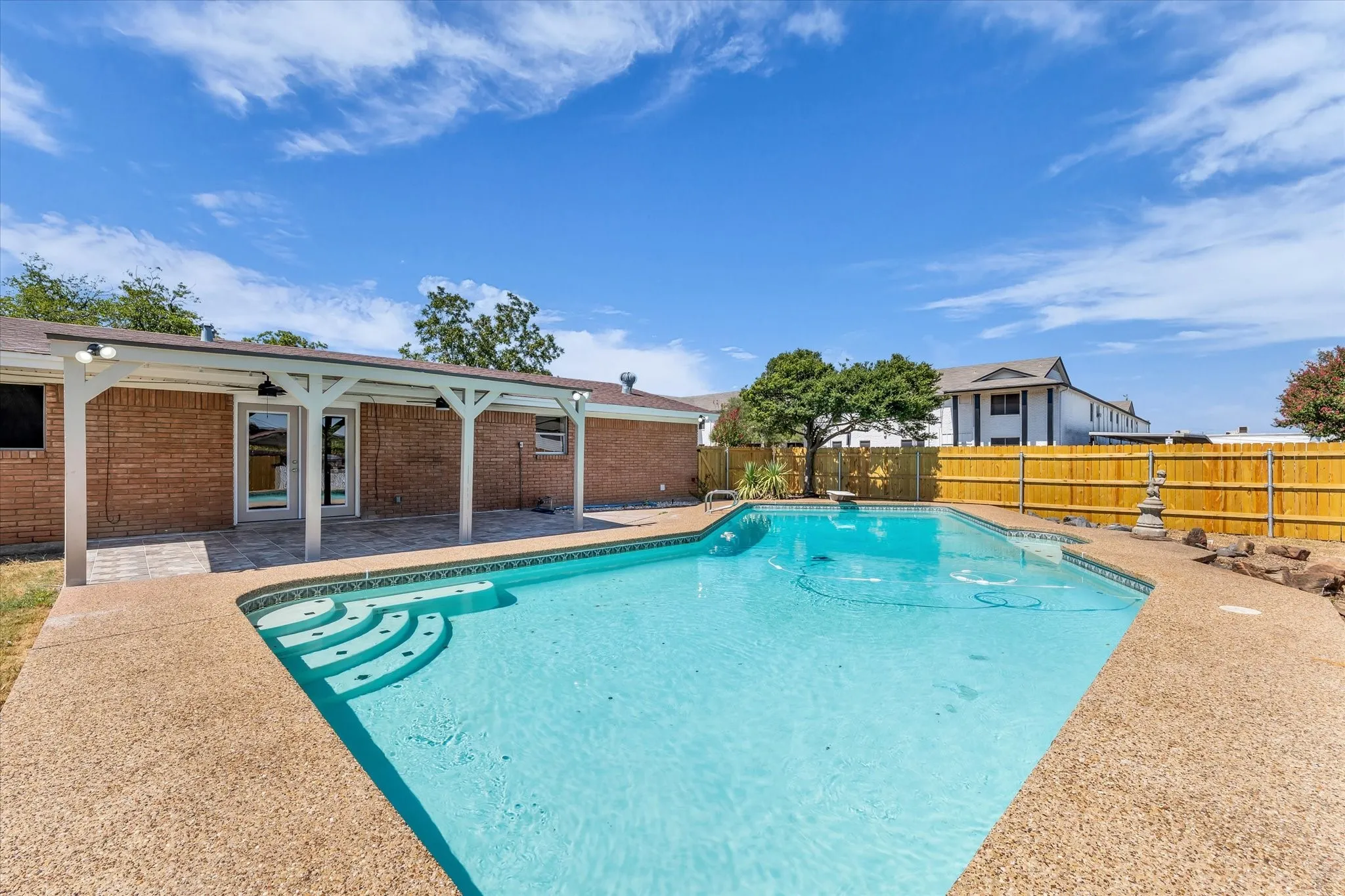 Expansive backyard featuring a swimming pool with integrated steps, a surrounding aggregate deck, and a covered patio with overhead lighting
