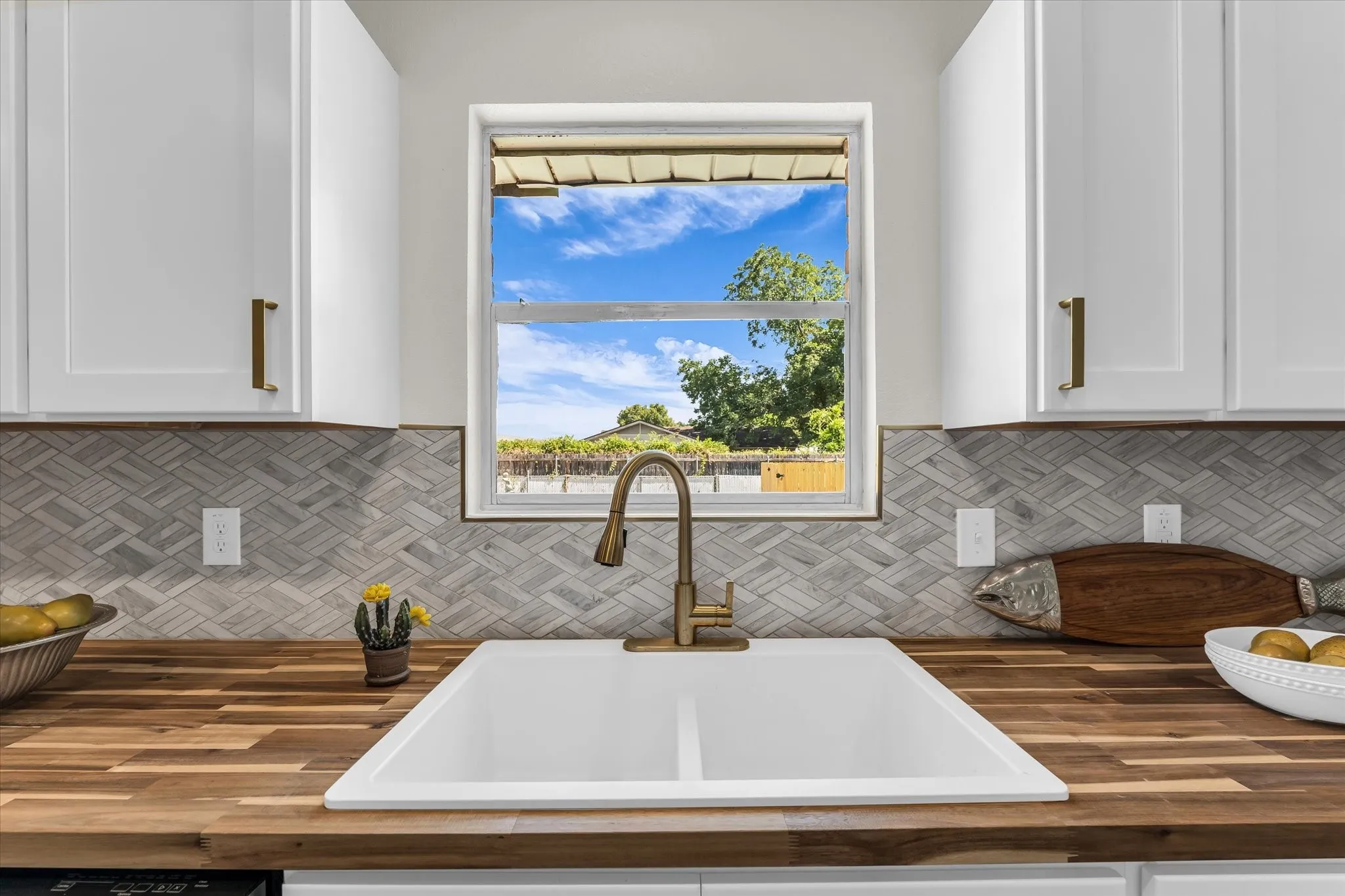 White cabinetry with brass hardware, wood-finish countertops, white double basin sink with brass faucet, and a herringbone pattern tile backsplash