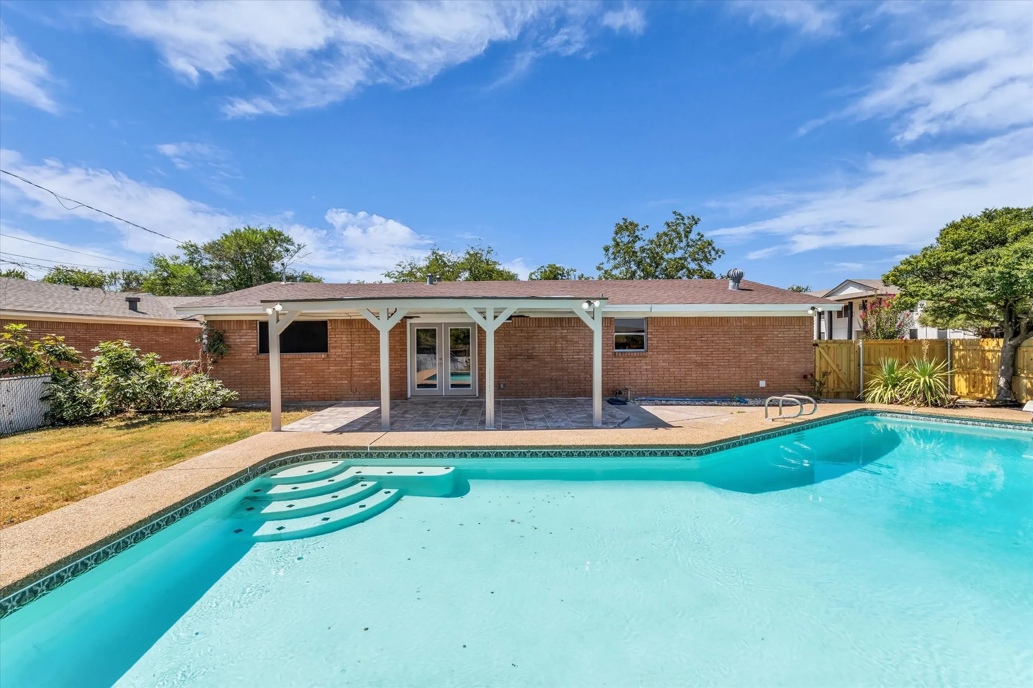 In-ground swimming pool with integrated steps, brick exterior, covered patio, double glass doors, and privacy fencing