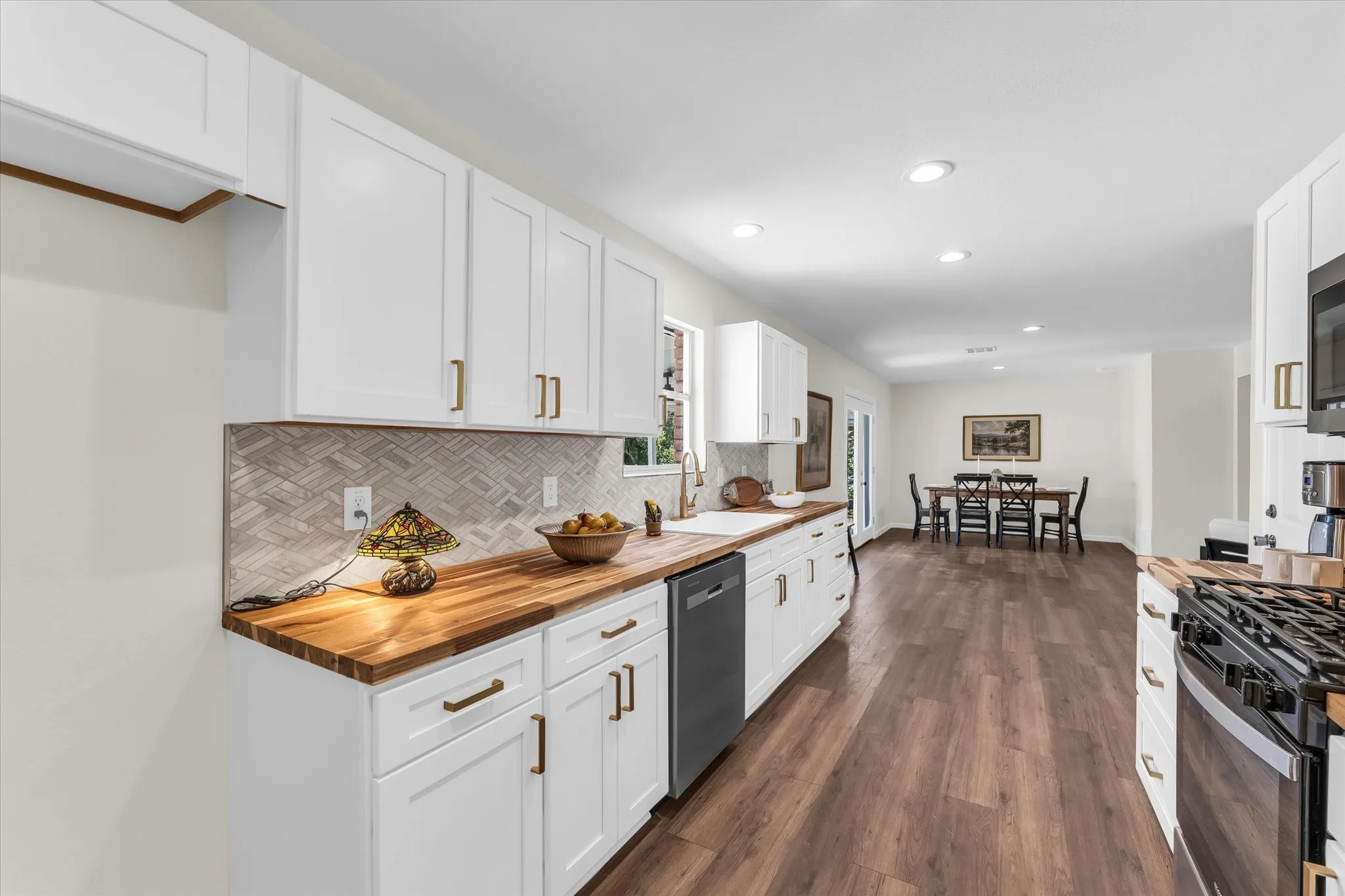 White shaker cabinetry with gold-finish hardware, wood-finish countertops, herringbone tile backsplash, and wood-finish flooring