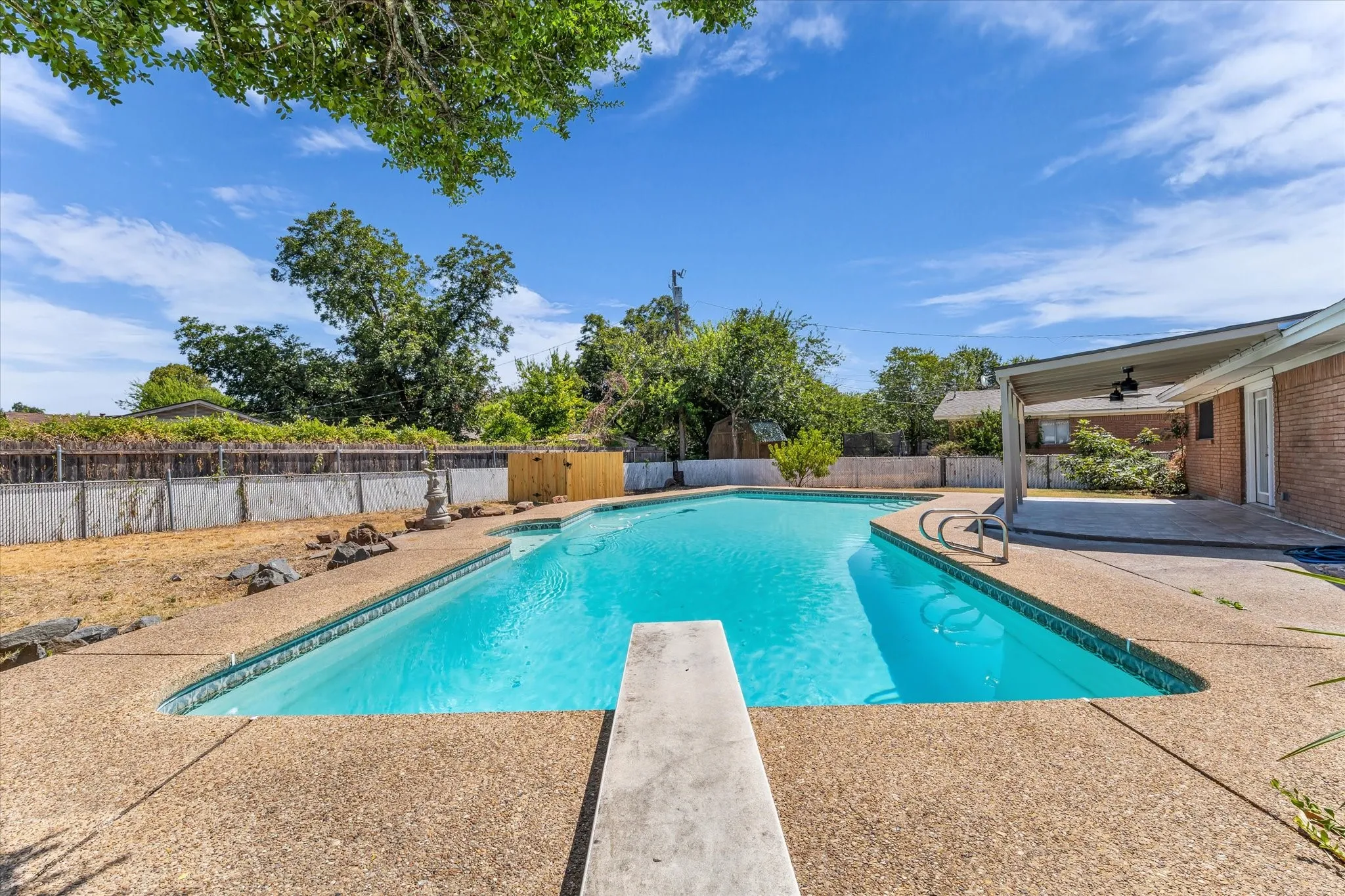 Outdoor swimming pool with a diving board, surrounding concrete decking, and covered patio with ceiling fans