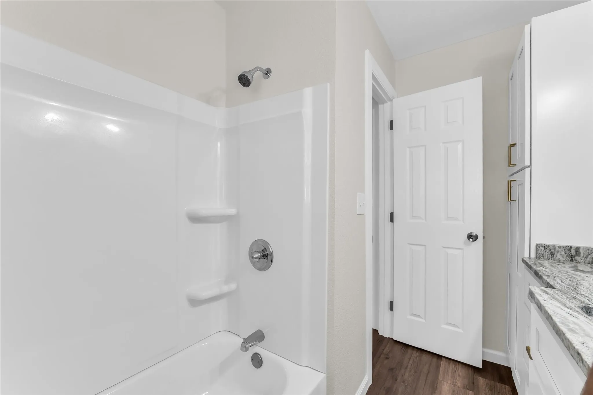 White acrylic tub and shower combination with chrome fixtures, wood-finish flooring, white paneled door, white cabinetry, and a stone-finish countertop