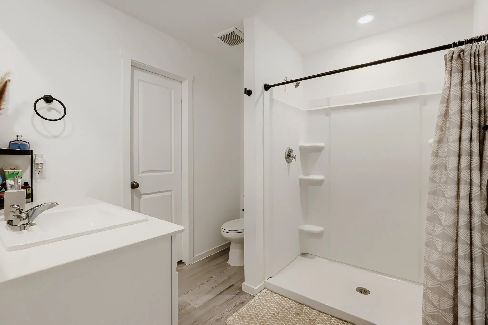 Bright bathroom featuring a single vanity with integrated sink, chrome faucet, white shower surround with built-in shelving, and wood-finish flooring