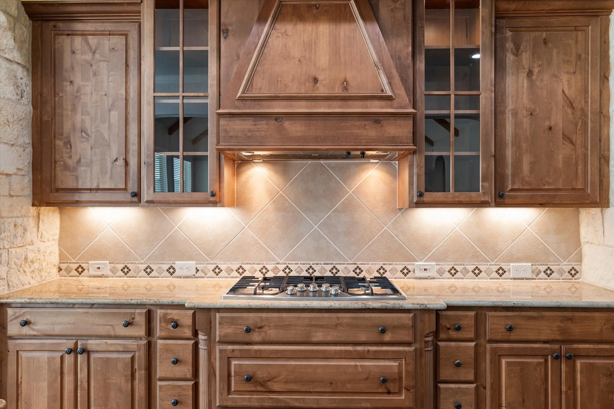 Wood cabinetry with glass-front accents, stone countertops, a tiled backsplash with decorative border, and a built-in gas cooktop