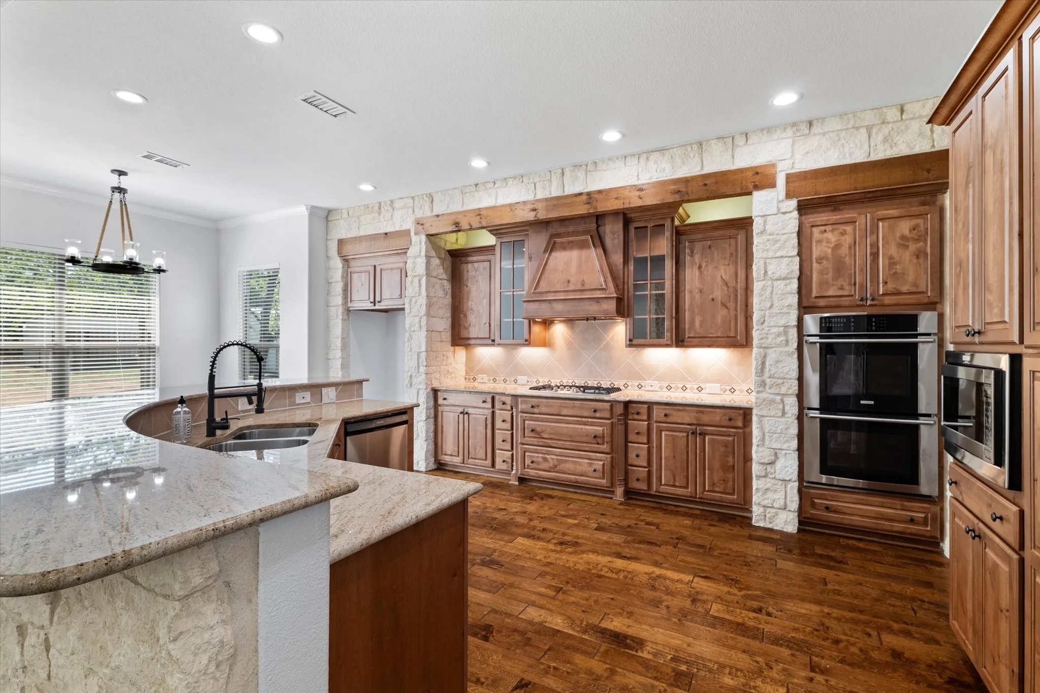 Spacious kitchen featuring a curved island with stone countertops, wood-finish flooring, custom wood cabinetry, and built-in stainless steel appliances