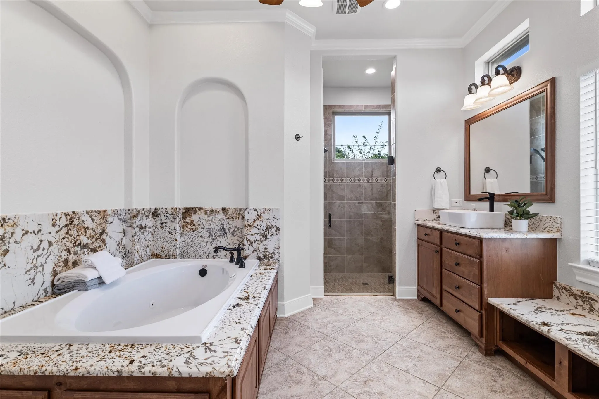 Soaking tub with stone surround, separate shower with tile detailing, wood-finish vanity with vessel sink, stone countertops, and tile flooring