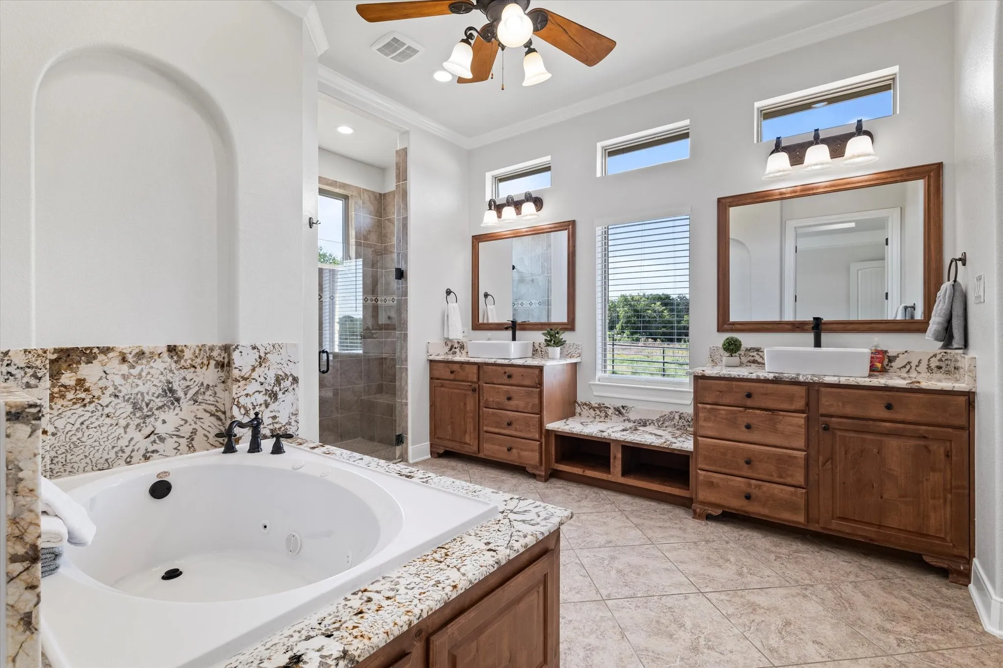 Built-in soaking tub with a stone surround, separate glass-enclosed shower, dual wood vanities with stone countertops, and tile flooring