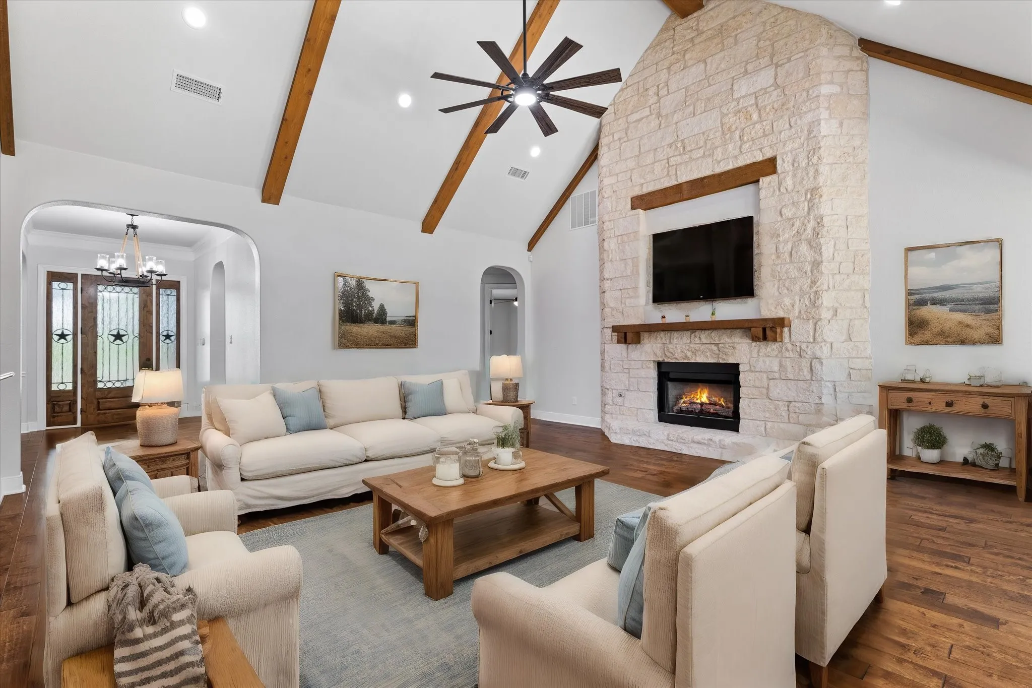 Vaulted ceiling with exposed wood beams, stone-clad fireplace, wood-finish flooring, recessed lighting, and a decorative front entry door with transom windows
