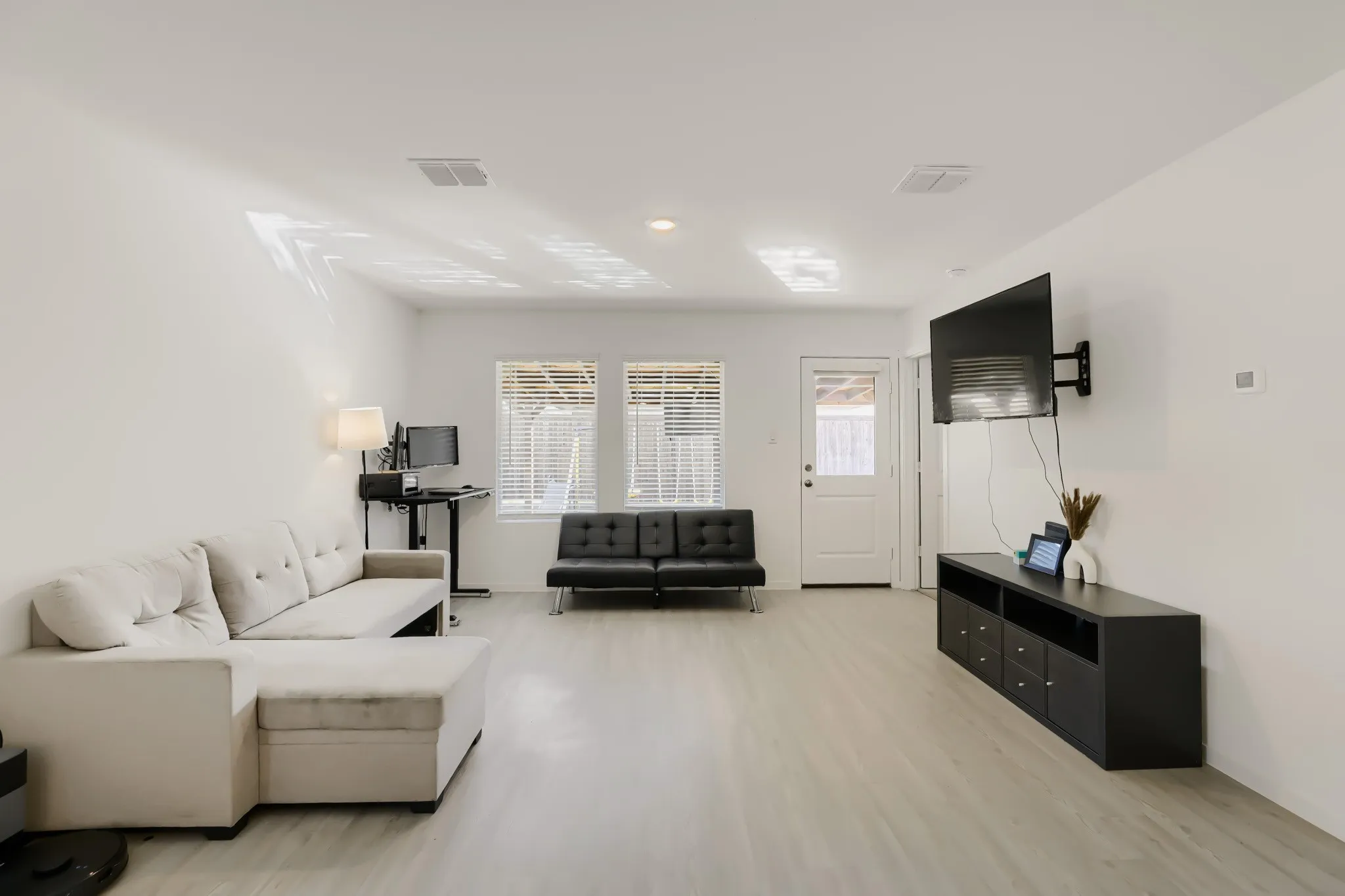 Spacious interior with light wood-finish flooring, white walls, multiple windows with blinds, and a white door providing exterior access