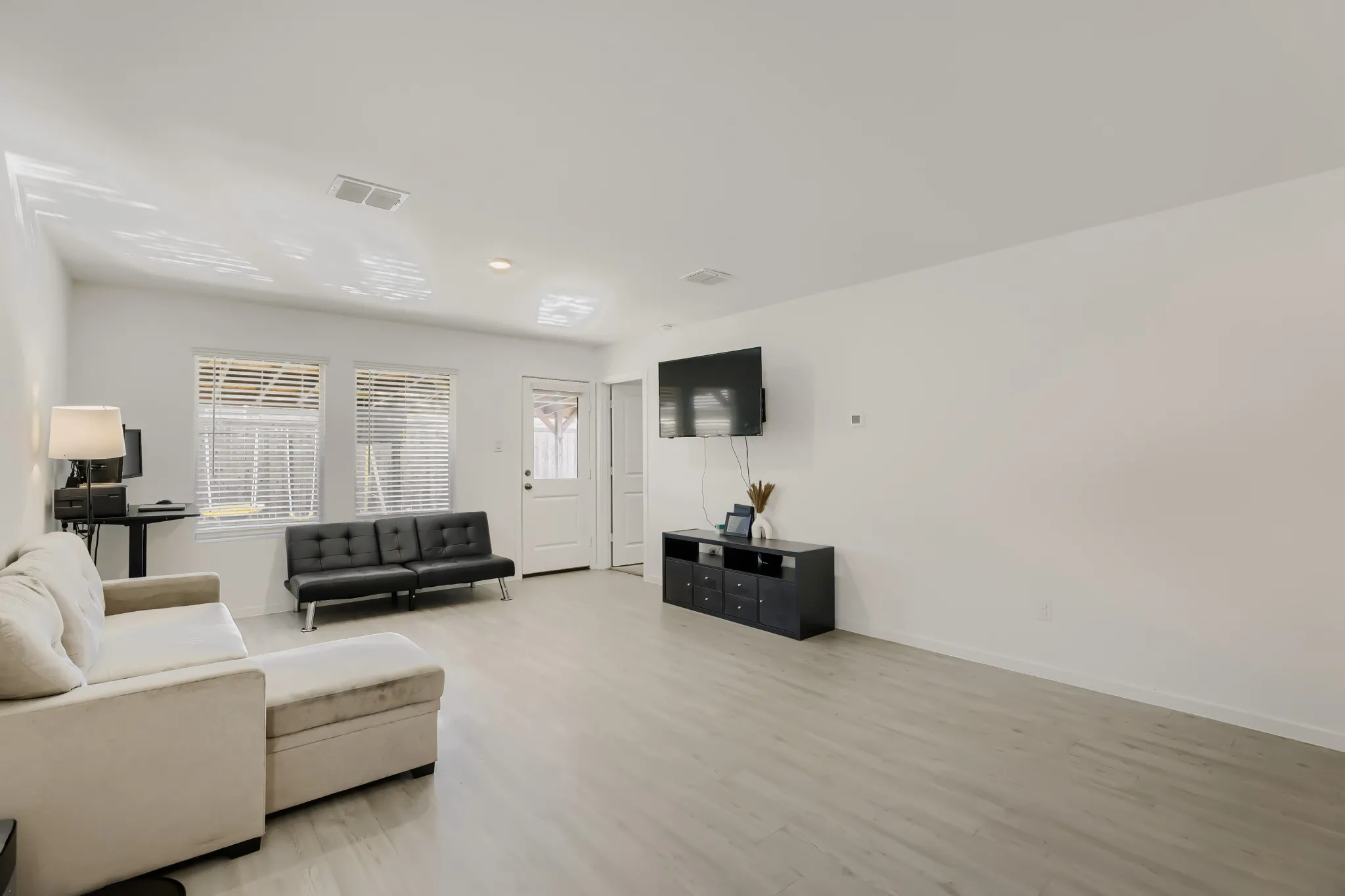 Open-concept living area with light wood-finish flooring, white walls, and multiple windows with blinds