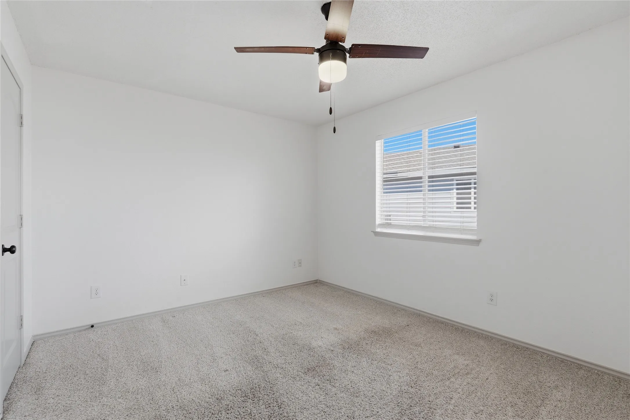 White interior walls, light-toned carpet, ceiling fan with integrated lighting, and a window with blinds