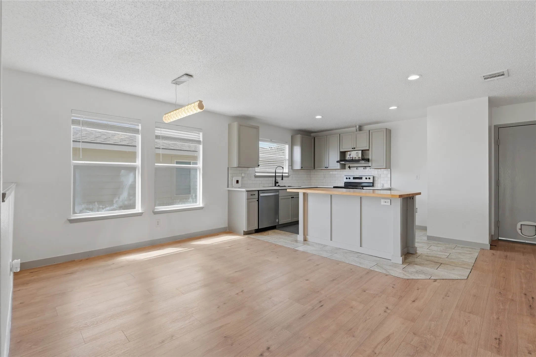 Open-concept living area with wood-finish flooring, a contemporary linear chandelier, and a kitchen featuring light gray cabinetry, stainless steel appliances, and a tiled backsplash