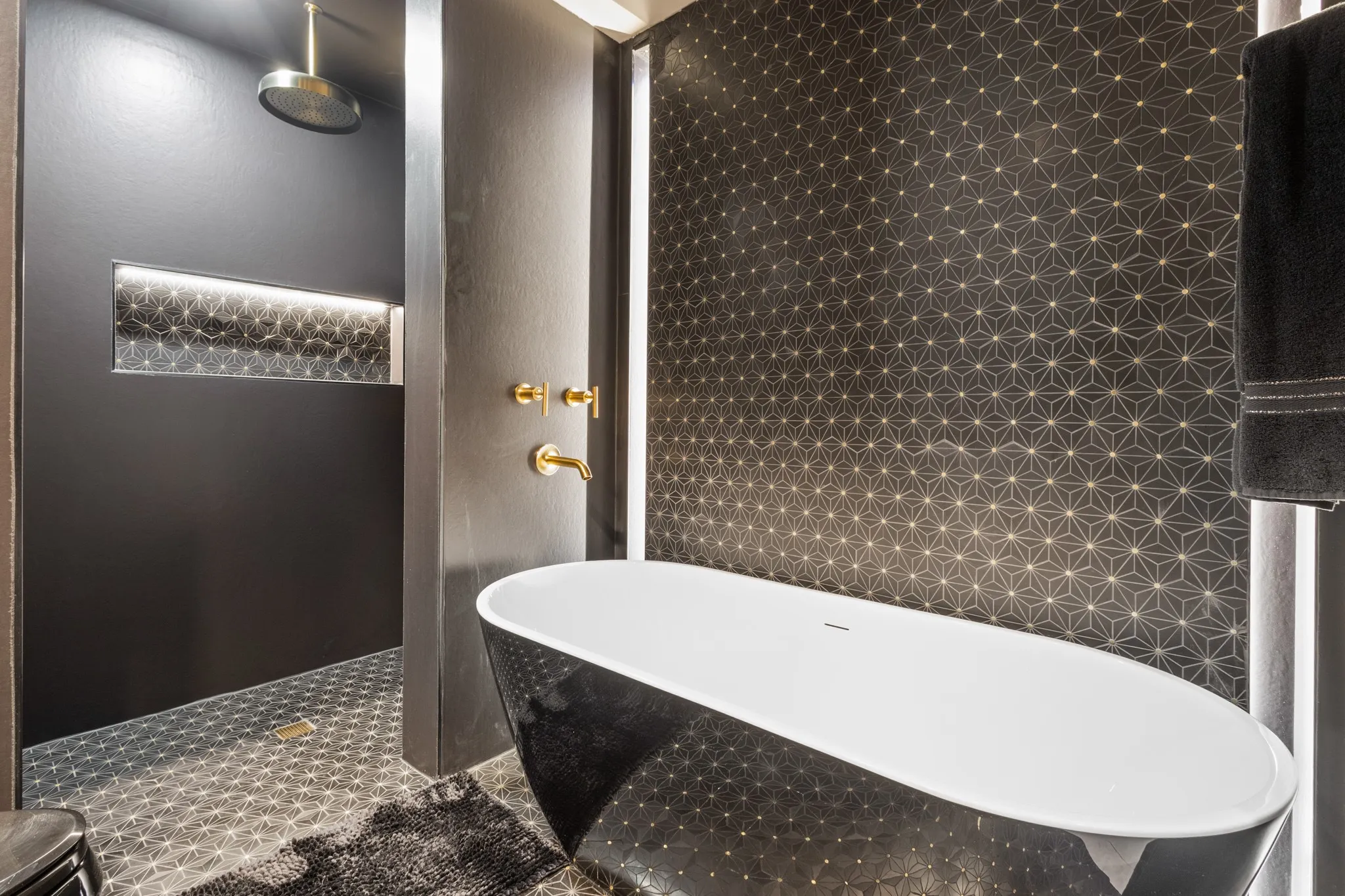 Spa-inspired primary bathroom featuring a freestanding oval soaker tub with integrated deck, geometric hexagonal tile work in warm bronze tones, rainfall showerhead, brass fixtures, and heated towel rack