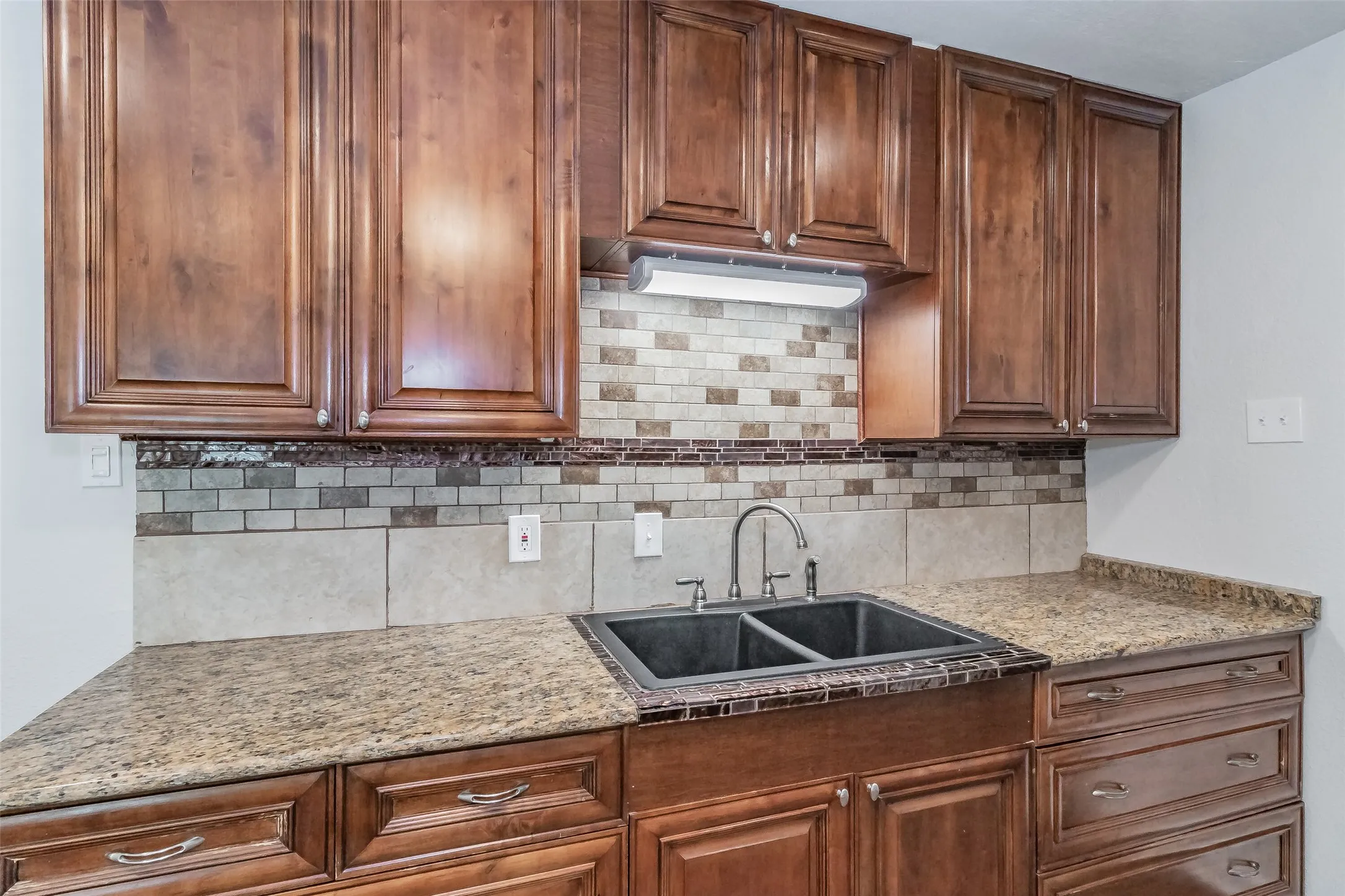 Wood-finish cabinetry with silver-tone hardware, textured tile backsplash, granite-style countertops, and a dual basin sink with a chrome faucet