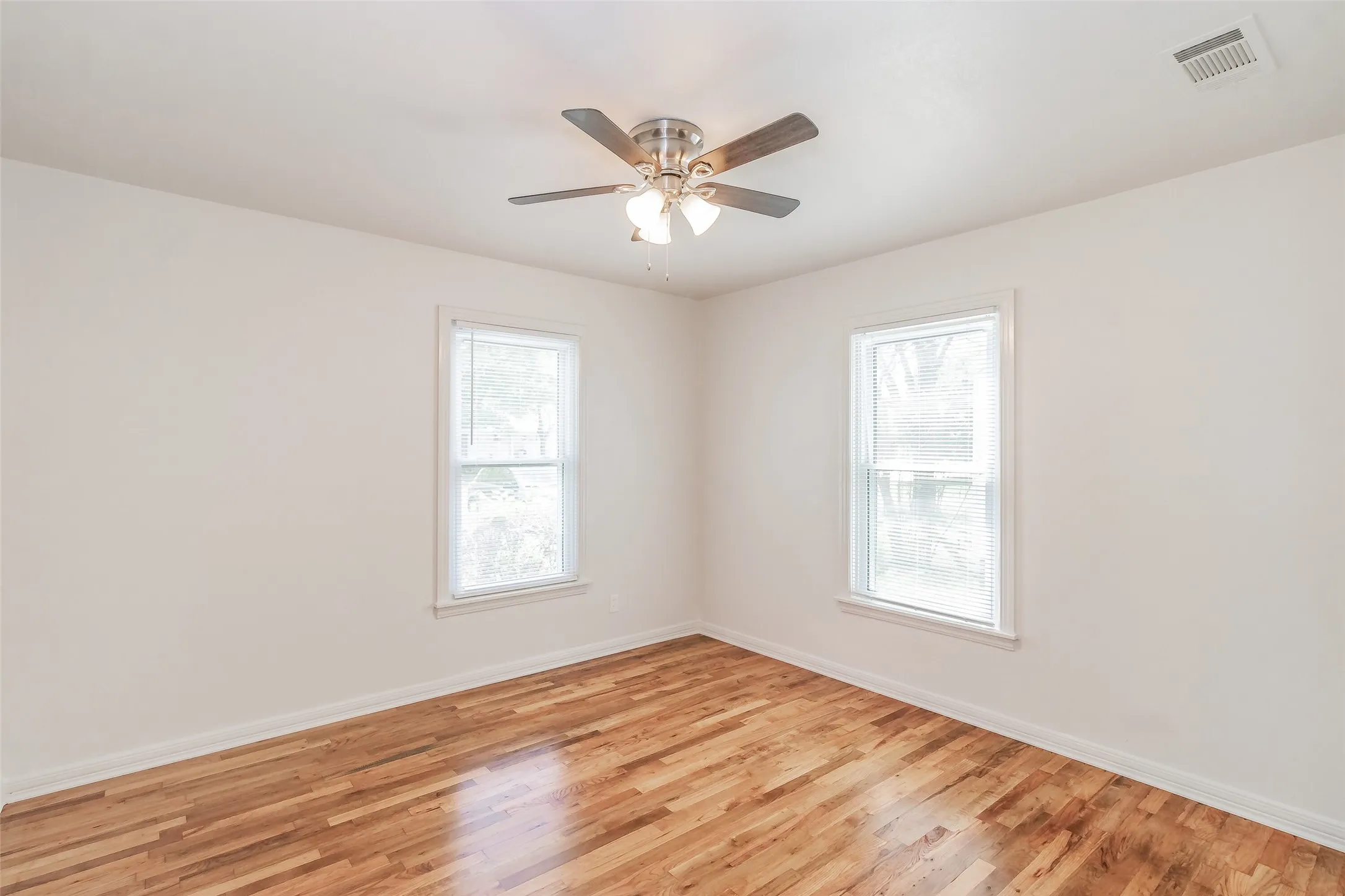 Wood-finish flooring, two large windows, ceiling fan with integrated lighting, and neutral wall paint