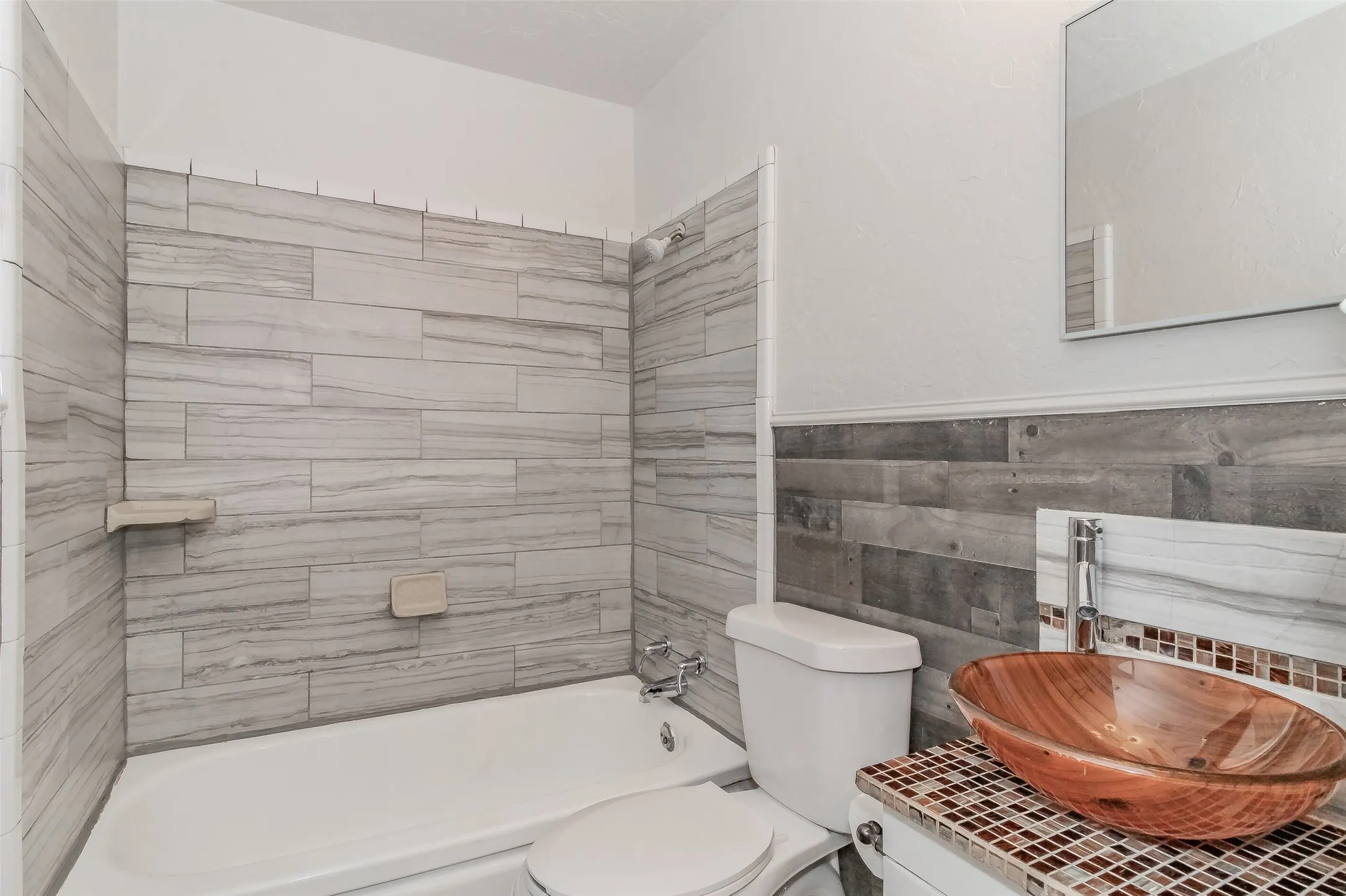 Integrated bathtub and shower with white subway tile surround, wood-finish accent wall, vessel sink, and wall-mounted mirror