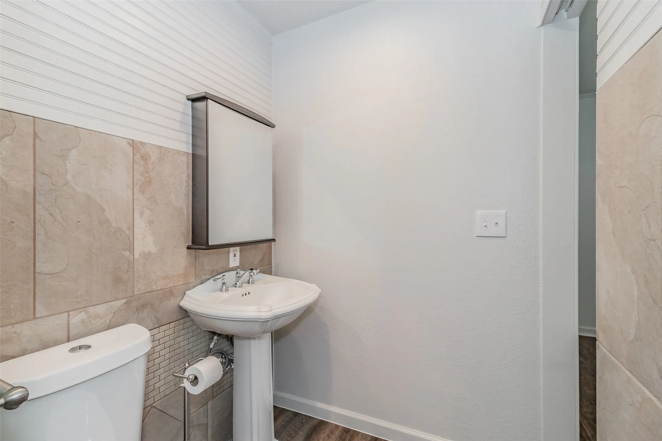 Pedestal sink with chrome fixtures, tiled wainscoting, white wall paneling, and wood-finish flooring