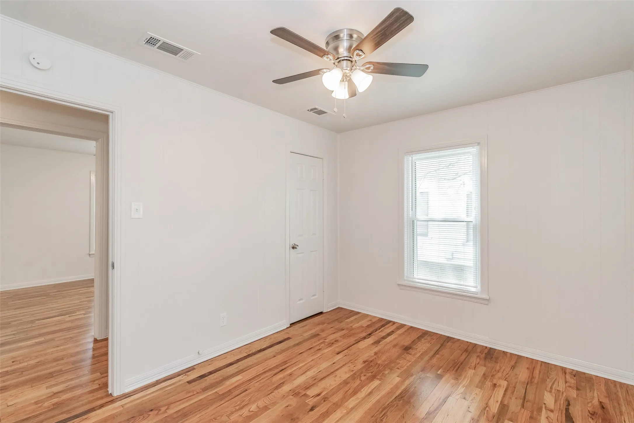 Wood-finish flooring, white painted walls, ceiling fan with light fixture, window with white trim, and white paneled door