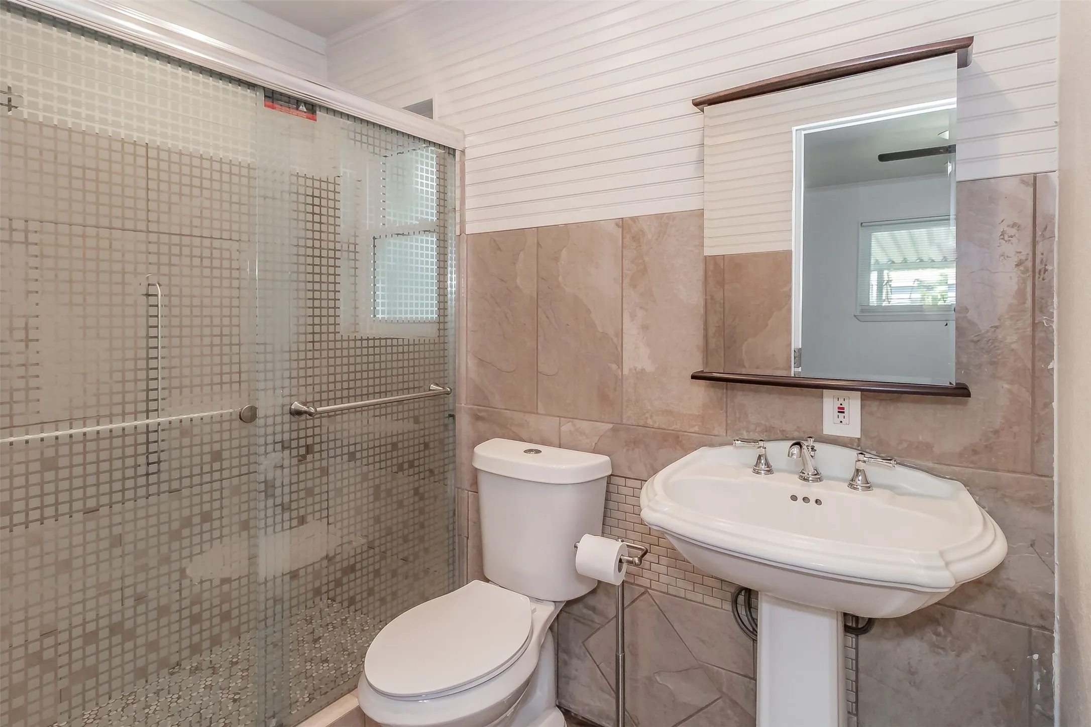 Pedestal sink with chrome fixtures, full mirror with wood trim, wall-mounted toilet, and a glass-enclosed shower featuring tile surround