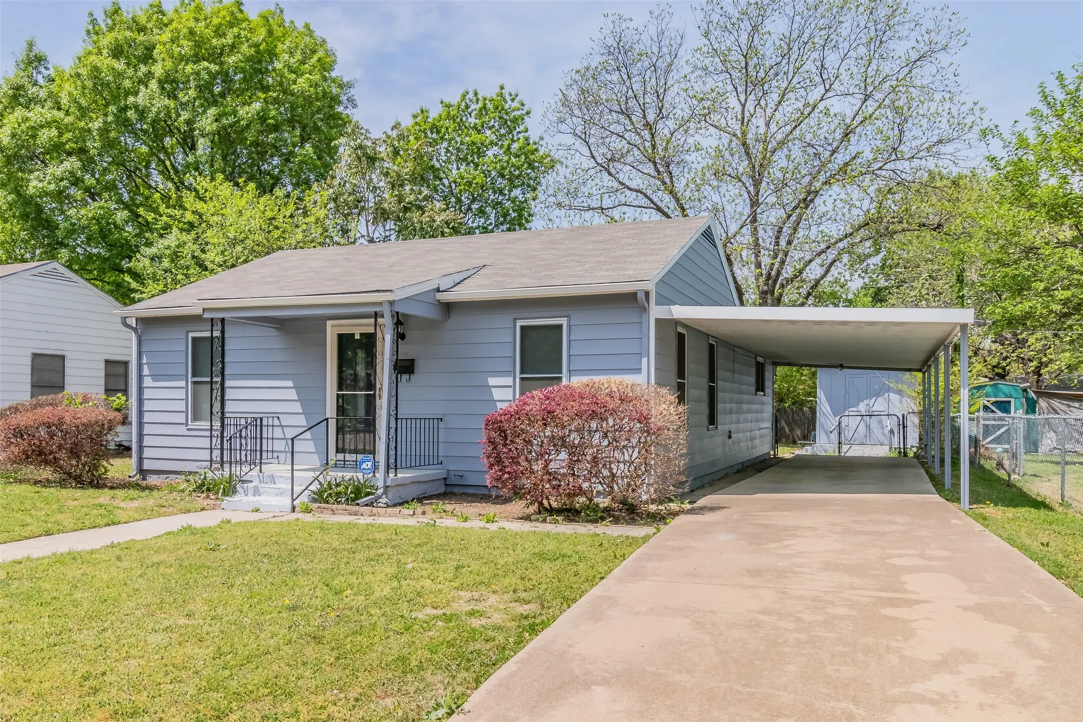 Light blue siding exterior with a covered front porch, concrete driveway, and a metal carport structure