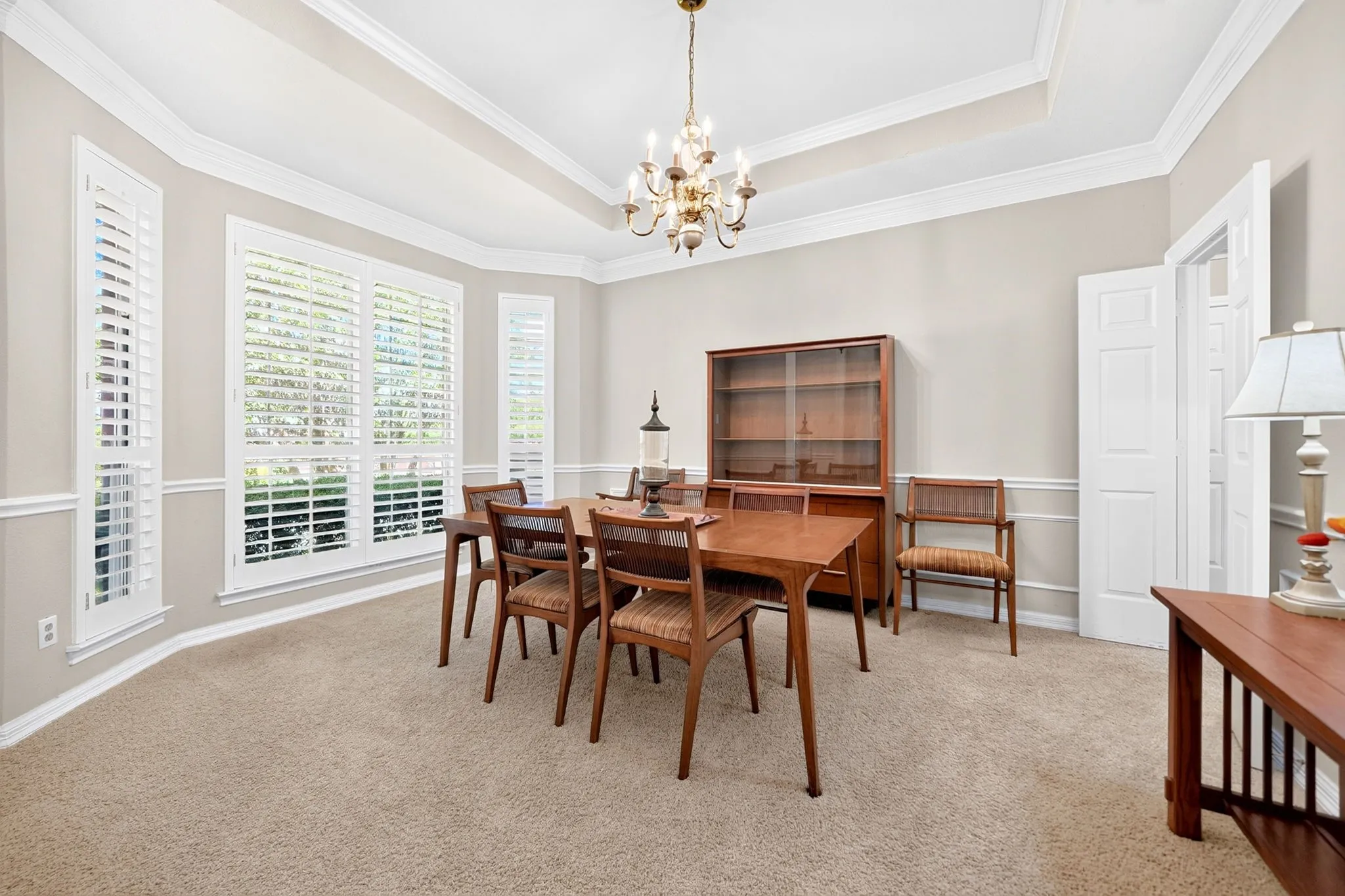 Tray ceiling with crown molding, bay window with plantation shutters, wainscoting, and carpeted flooring