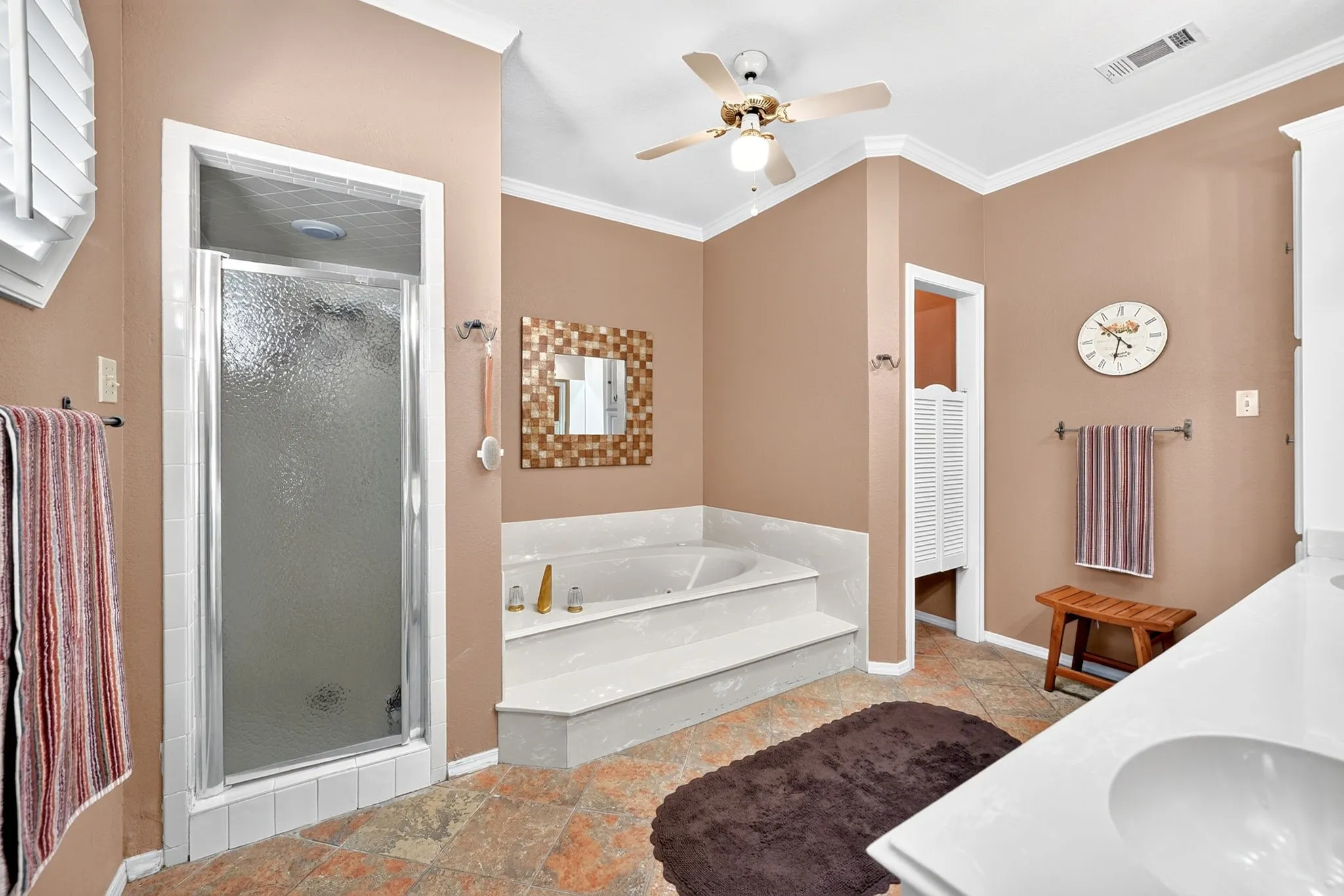 Soaking tub with tiled surround, walk-in shower with textured glass door, dual vanity with integrated sinks, and a ceiling fan