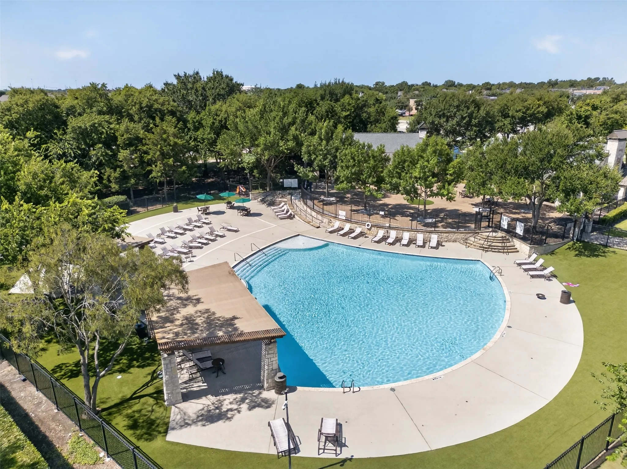 Expansive outdoor pool with multiple entry steps, surrounding hardscaping, a stone-clad cabana, and perimeter fencing