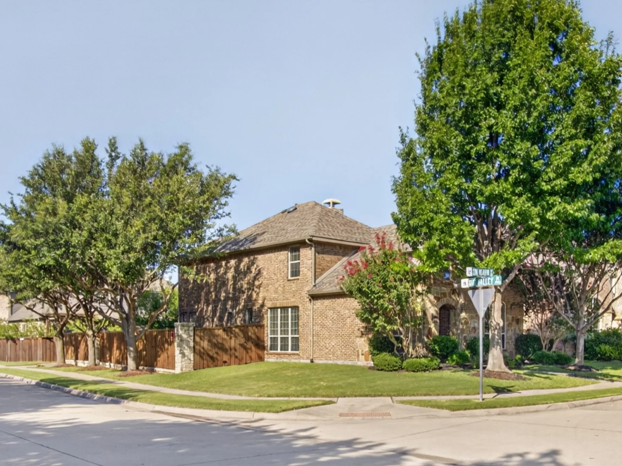 Brick exterior with a shingled roof, mature trees, and a wood privacy fence with stone pillars