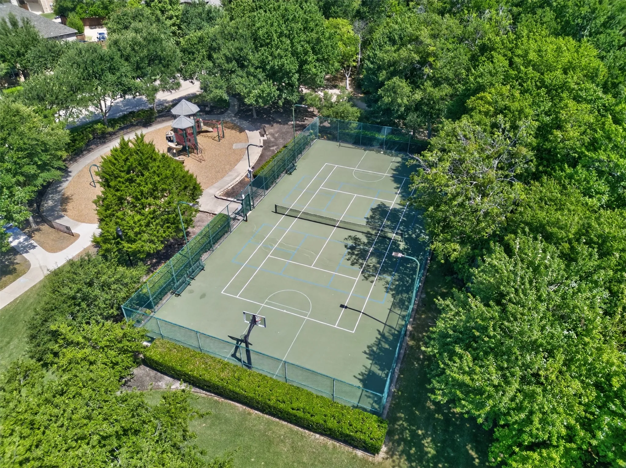 Tennis court with basketball hoop, playground area with wood chip surfacing, paved walking paths, and mature tree landscaping