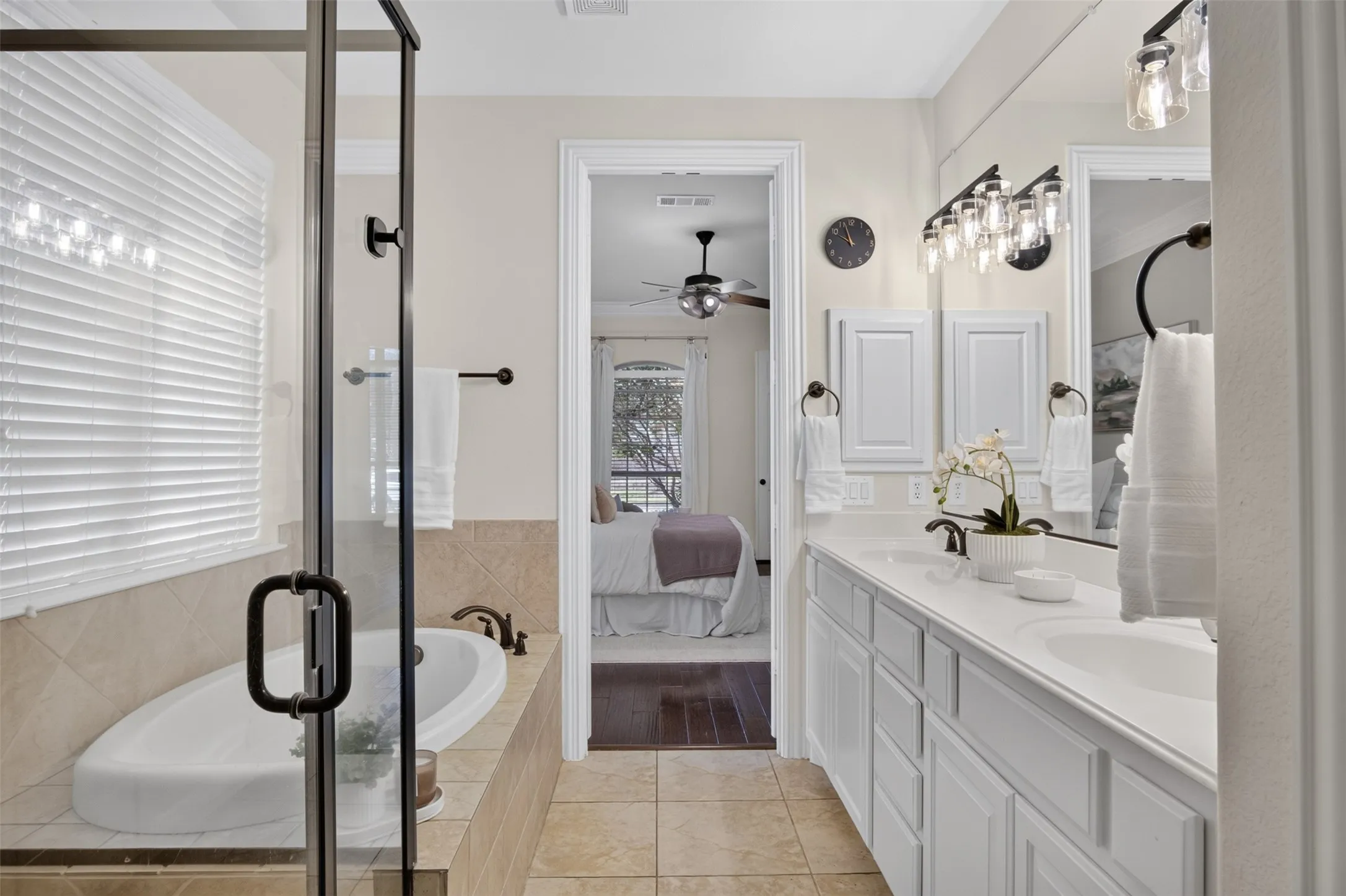 Dual vanity with white cabinetry and integrated sinks, a tiled soaking tub with dark bronze fixtures, a separate glass-enclosed shower, and tile flooring