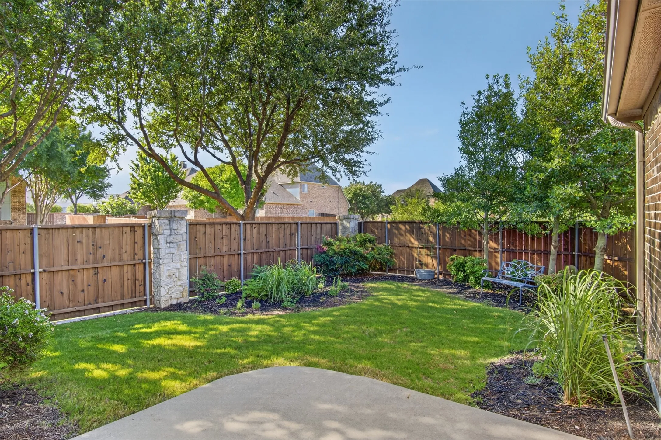 Private backyard featuring a concrete patio, wood fencing with stone pillars, mature trees, and landscaped garden beds